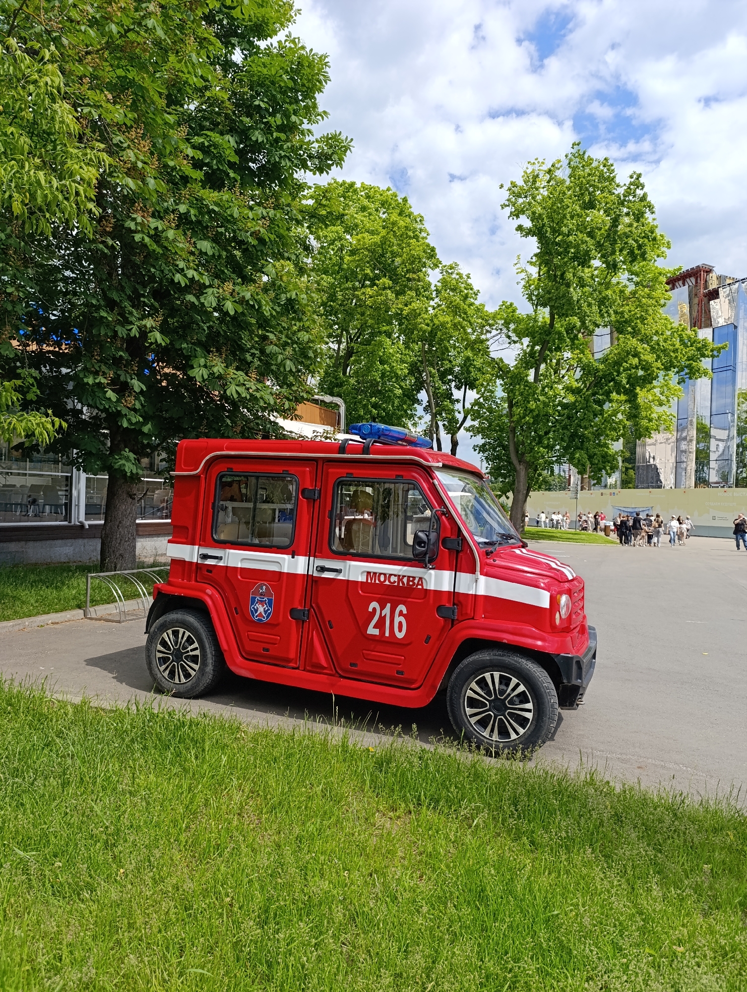 A red, compact fire truck is parked on a paved surface, serving as the central subject of this daytime outdoor scene in Moscow, Russia. The vehicle is bright red with two white horizontal stripes along its side and a blue light bar mounted on the roof. On the driver's side door, a circular emblem featuring a coat of arms is visible, accompanied by the word "МОСКВА" (MOSCOW) in white Cyrillic letters and the prominent number "216". The truck's wheels have black and silver multi-spoke rims.
In the foreground, a vibrant stretch of green grass leads up to the paved area. Lush, green-leaved trees, possibly chestnuts with developing fruits, frame the scene, providing shade and indicating a spring or summer season. Behind the trees on the right, a modern building with reflective glass and metallic facades stands under a partly cloudy sky, which shows patches of blue.
In the mid-ground, a group of people can be seen strolling or gathering on the paved area, suggesting a public park or square. A silver bicycle rack is visible near a building on the left. The overall impression is one of a bright, clear day.