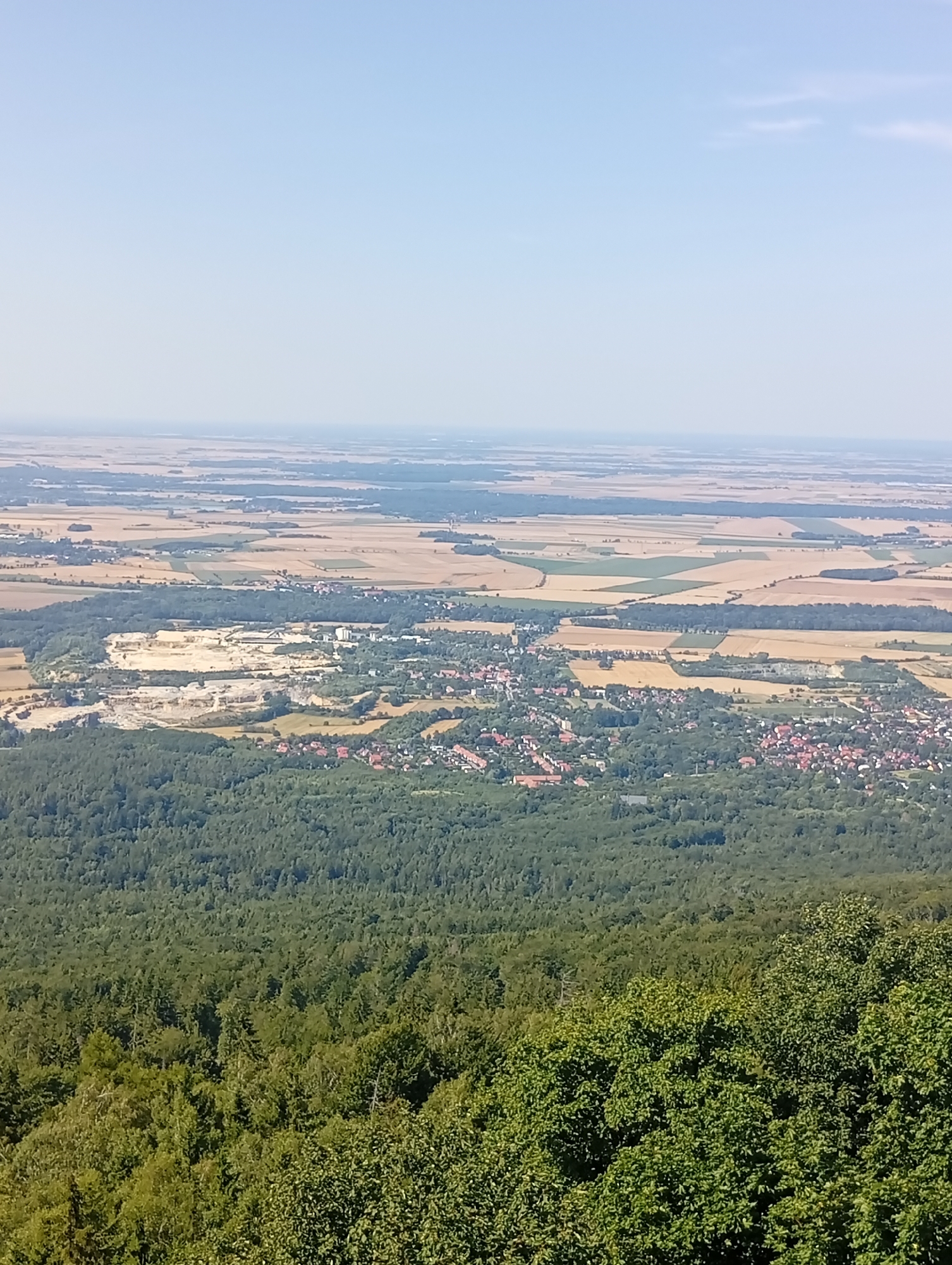 This is a landscape photograph taken from a high vantage point overlooking the city of Sobótka, Poland. In the foreground, a dense forest of green trees fills the bottom portion of the image. Beyond the forest, the city of Sobótka is visible, characterized by a cluster of red-tiled roofs and buildings nestled amongst more trees. Further in the midground, a large, open quarry with pale, layered rock formations is prominent. The background reveals a vast expanse of agricultural fields in various shades of brown and green, stretching towards a hazy horizon under a pale blue sky. The overall scene depicts a rural landscape with urban and industrial elements integrated within it.