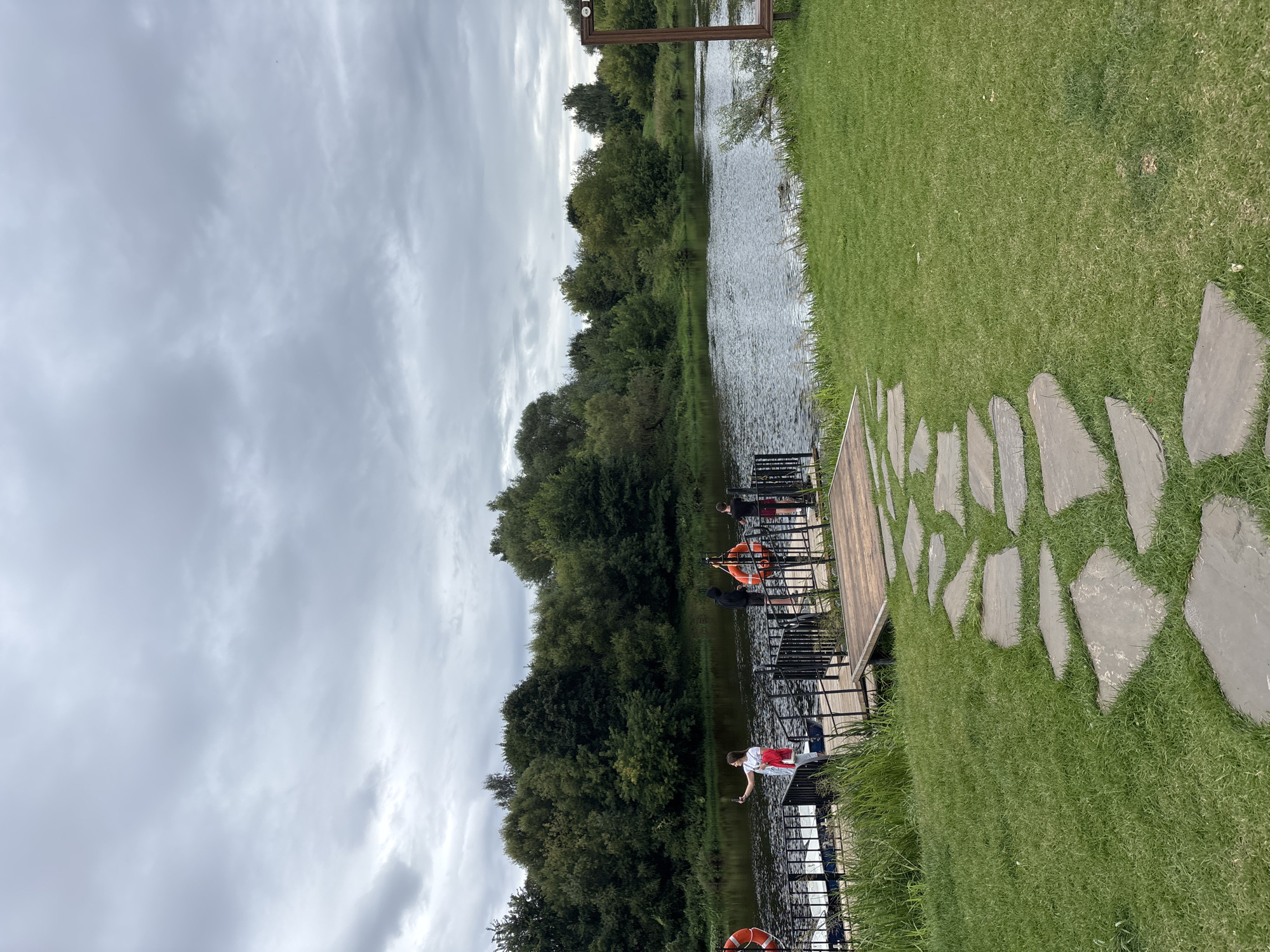 The image depicts a cloudy day by a river, likely Zvenigorod, Russia, given the context provided. In the foreground, a grassy area features a winding path made of large, flat stones. To the left, a wooden dock extends into the dark, rippling water, bordered by lush green trees and vegetation on the far bank. On the dock, several people are visible. One woman in a white shirt and red jacket is taking a selfie with her phone. Two men, one in a black hooded sweatshirt and another in a black t-shirt and red shorts, are standing near orange life preservers attached to the dock's railing. A third person, partially obscured, is also on the dock. The overall atmosphere is tranquil, with a hint of recreational activity.