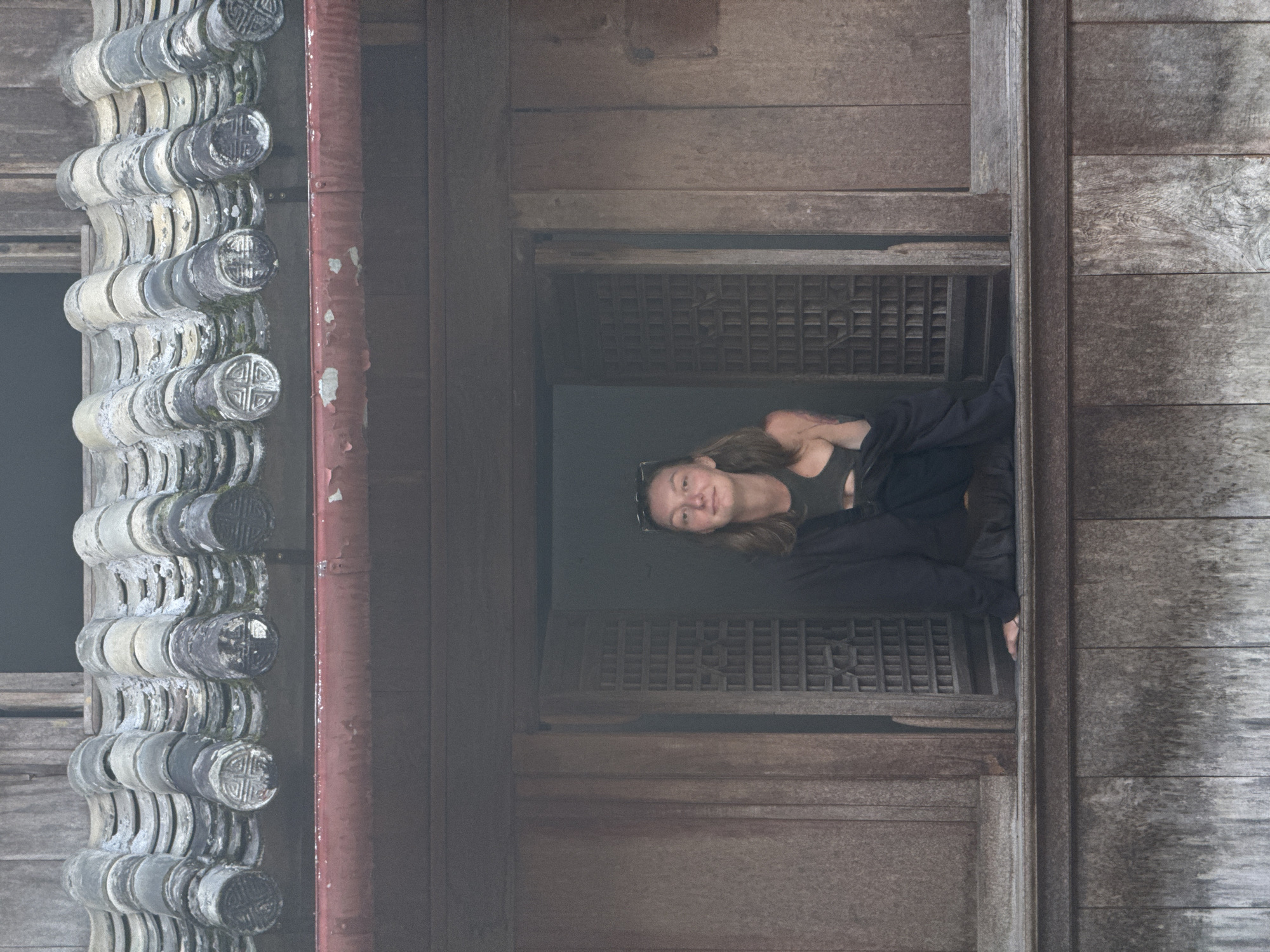The image shows a woman looking out of a window in a building in Sà Phìn Commune, Vietnam. The building appears to be made of wood and is weathered, suggesting age. The window has a latticework pattern. The woman has fair skin and is wearing dark clothing. Exterior to the window is a column of grey roof tiles. A red pipe runs vertically along the wooden facade next to the tile. The image is taken at an angle.