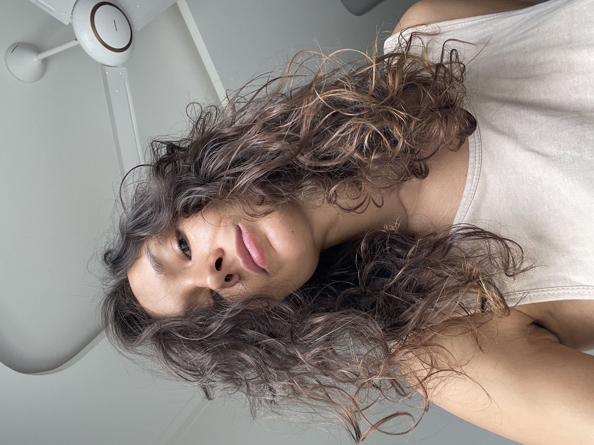 A person is lying down and looking up at the camera, positioned in a location with a ceiling fan, likely indoors. The person has curly, dark brown hair with some lighter brown highlights. They are wearing a white tank top. The ceiling and walls appear to be white. The location is unspecified but implied to be in the city of Mũi Né, Vietnam.