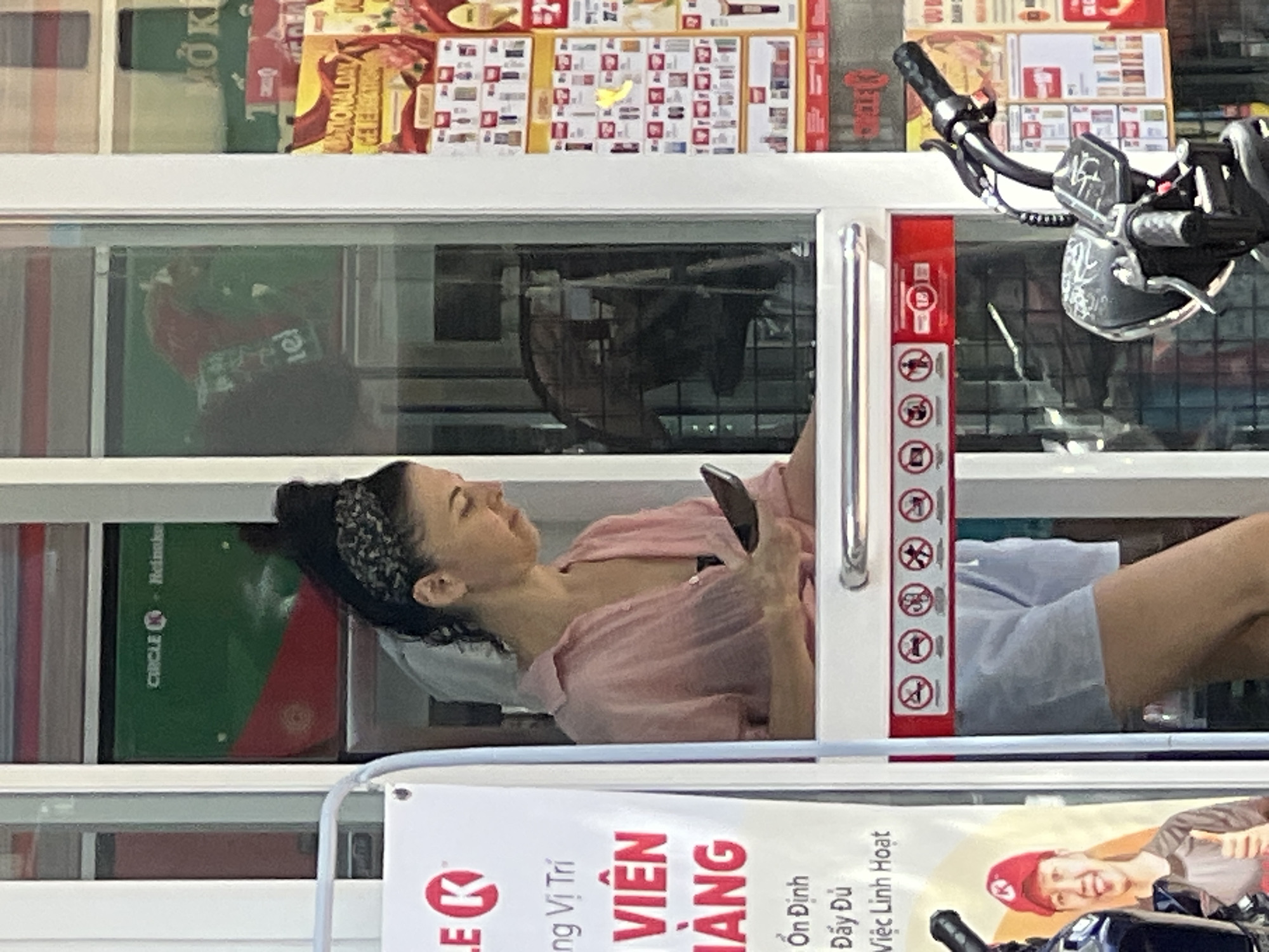A woman wearing a light pink collared shirt and light blue shorts stands at a window, looking down at her phone. She has dark hair pulled up and is wearing a patterned headband. Behind her, a refrigerator unit with "CIRCLE K Heineken" branding is visible. To the left of the woman is a white sign with Vietnamese text and the Circle K logo. To the right, a motorcycle is parked. The scene appears to be at a Circle K convenience store in Nha Trang, Vietnam, given the Vietnamese signage.