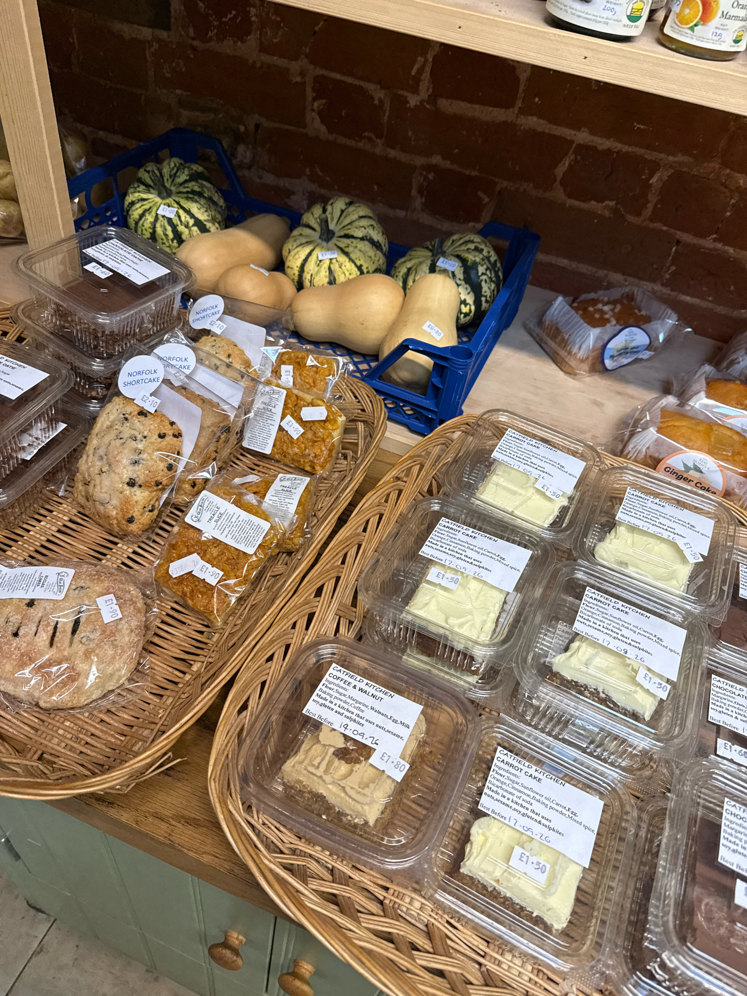 The image shows a display of baked goods and produce in what appears to be a shop or market stall. In the background, a blue plastic crate contains several small gourds and squashes, some of which have price tags indicating "£1.50". To the left, a wooden shelf holds jars of preserves, one of which is labeled with an orange slice.
In the foreground, two woven baskets are filled with various packaged food items. The left basket contains a large round loaf of bread with visible scoring and what appears to be dried fruit or nuts embedded in its crust, priced at "£1.50". Next to it are several packaged cookies or shortbread. The right basket is predominantly filled with individually packaged slices of cake. Several of these slices are labeled "CATFIELD KITCHEN CARROT CAKE" with ingredients and "Best Before 17.09.26" visible, priced at "£1.50" each. Other packaged items in this basket include what look like traybakes, with labels indicating "TREACLE" and prices of "£1.90". Further to the right, another packaged cake slice labeled "GINGER CAKE" is visible. Below the cakes, a clear plastic container holds a portion of coffee and walnut cake.
The overall environment suggests a food retail setting with a rustic or artisanal feel, characterized by the woven baskets and wooden elements. The items are neatly arranged for sale. The presence of price tags and "Best Before" dates indicates a commercial transaction context. The lighting appears to be ambient, possibly from overhead sources within the establishment.