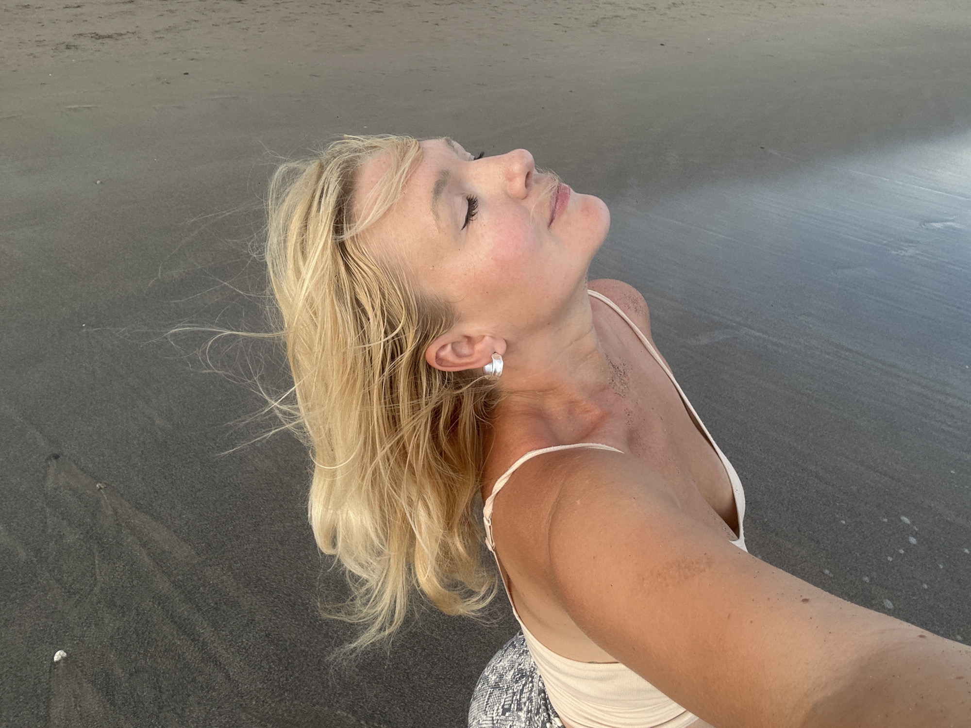 A blonde woman is standing on a dark sand beach in North Kuta, Indonesia. She is looking up with her eyes closed and her face tilted back. Her hair is blowing in the wind. She is wearing a light-colored tank top and patterned pants. The beach is wet and there are some small shells scattered on the sand. The water is visible in the background.
