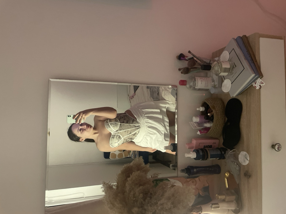 A female subject is depicted taking a mirror selfie in an indoor setting, identified as Соболиха, Russia. The subject, an adult female with dark hair, wears a strapless, patterned corset-style top and a white, ruffled mini-skirt. She holds a light green smartphone in her right hand, angled to capture her reflection, and displays a slight smile. A faint purplish residue is visible on her face, consistent with a facial mask.
The mirror, rectangular and unframed, reflects the subject and the room behind her. To the immediate left, a large bundle of dried ornamental grass is arranged in a vase on a light wood vanity table. The table surface is cluttered with various personal care and cosmetic items, including multiple bottles of lotions and sprays, a small woven basket holding more products, a black eye mask, and a gold-colored tray. On the right side of the table, a bottle of Bioderma micellar water, a glass containing several makeup brushes, a perfume bottle, and a stack of three books (with visible titles "PRADA" and "DIOR") are present. White cotton pads are scattered on the table.
In the background reflected in the mirror, a white-framed window with a light curtain is visible to the far left. A chair with a patterned upholstered back (cream and olive tones) is positioned behind the subject's reflection. Further back, a bed with light-colored bedding can be discerned. The room walls are plain and light-colored, possibly pale pink or beige. The lighting is soft and even, suggesting indoor artificial illumination.