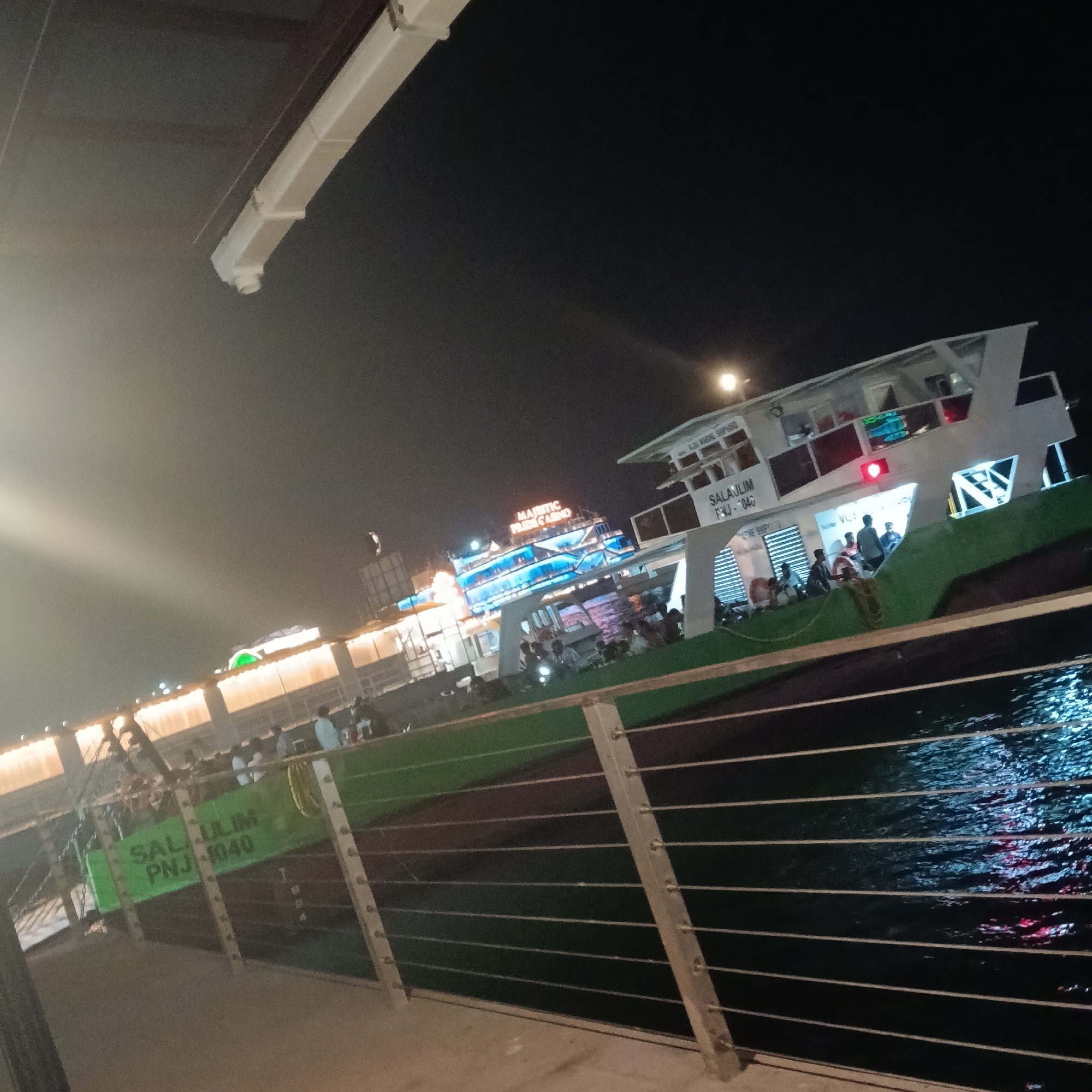 The image, taken at night, features a body of water with light reflections, a metal railing in the foreground, and two vessels docked in the background. The vessel closer to the foreground is labeled "SALAULIM PNJ-1040" and "SALAULIM FIJ-1040." The other vessel is identified as "MAJESTIC PRIDE CASINO." People are on board both vessels. The location is in the city of Panaji, India.