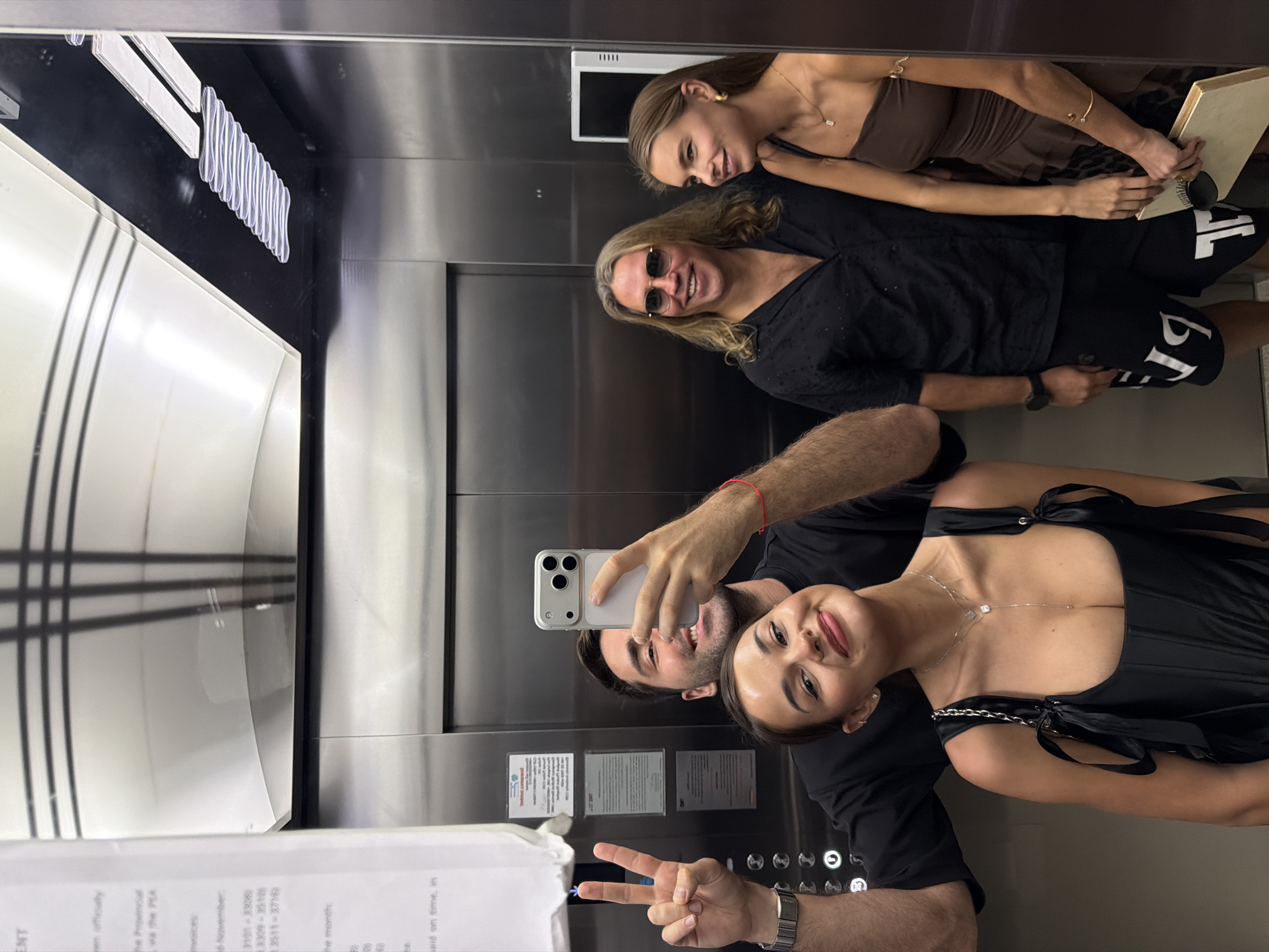 Here is a description of the image:
The image depicts four people in an elevator. A couple takes a selfie in the foreground. The woman is wearing a black dress with ribbon detailing and a necklace. The man is holding the phone. In the mirror's reflection, we see a woman in sunglasses and another woman in a brown dress. Part of a document or sign is visible at the bottom left of the image. The elevator has stainless steel walls. Based on textual elements in the document, the image may be taken in the city of Choeng Thale, Thailand.