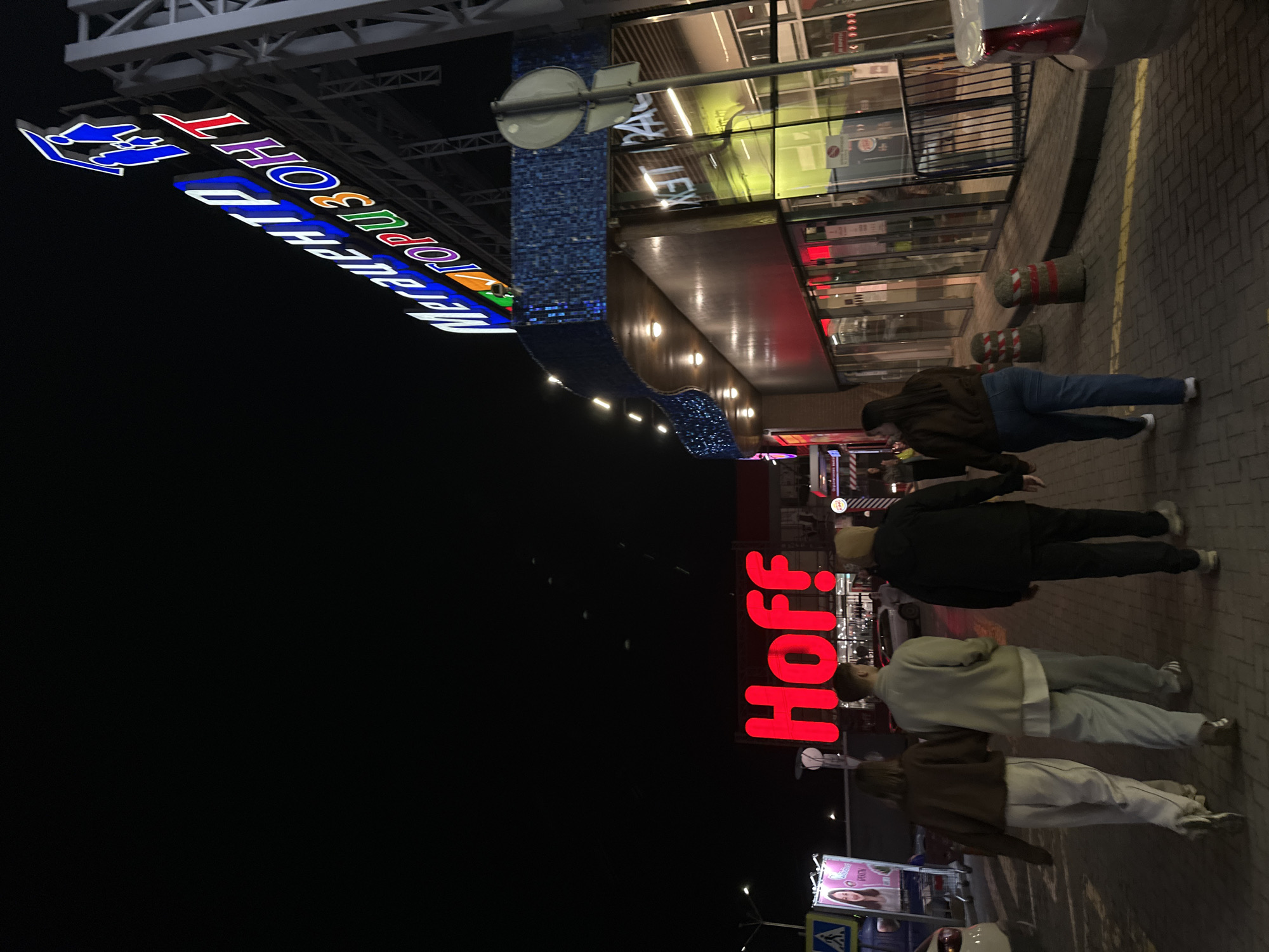 The image shows a nighttime scene outside a building in Rostov-on-Don, Russia. The building is lit with bright signs, including "Hoff" in large red letters and "Мегацентр Горизонт" (Megacenter Horizon) in blue and other colors. The storefront has glass windows with reflections.
Four people are walking on the sidewalk in front of the building. They appear to be dressed in casual clothing. One is wearing a brown jacket and jeans, another has a black jacket, and the last two wear lighter colored jackets.
The sidewalk is paved with gray bricks. There are red and white striped bollards near the building. A street sign with a pedestrian crossing symbol is visible in the lower part of the image, and a vehicle's taillight can be seen. The environment suggests a commercial area during evening or night.