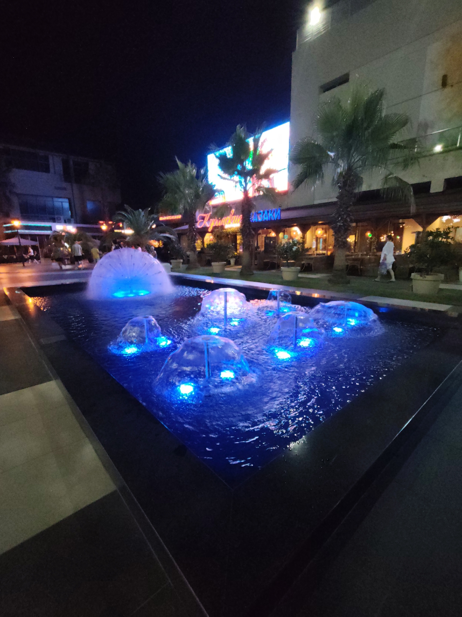 At night, a modern fountain with blue lights is located in a city square in Sochi, Russia. The fountain is rectangular and filled with water, with several jets of water shooting up in various shapes. Some of the jets are illuminated with blue LEDs, creating a vibrant display. Palm trees and buildings line the square, and several people are walking in the background. The scene is well-lit, and the water from the fountain is reflecting the lights, creating a magical atmosphere.