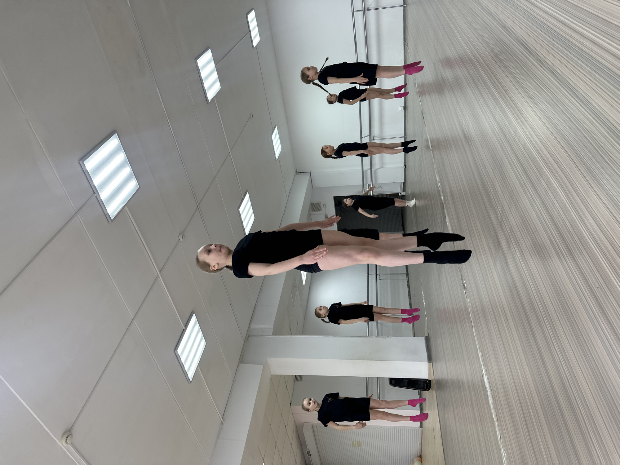 The image shows an indoor dance studio. Multiple girls are present, dressed in black leotards and socks (some pink, some black). They appear to be practicing dance or ballet. The studio has a ballet barre along one wall, and the floor is light-colored with linear patterns. The walls are white, and rectangular ceiling lights are visible. The reflection of the dancers is visible in a mirror. It is not possible to confirm if this is Votkinsk, Russia.