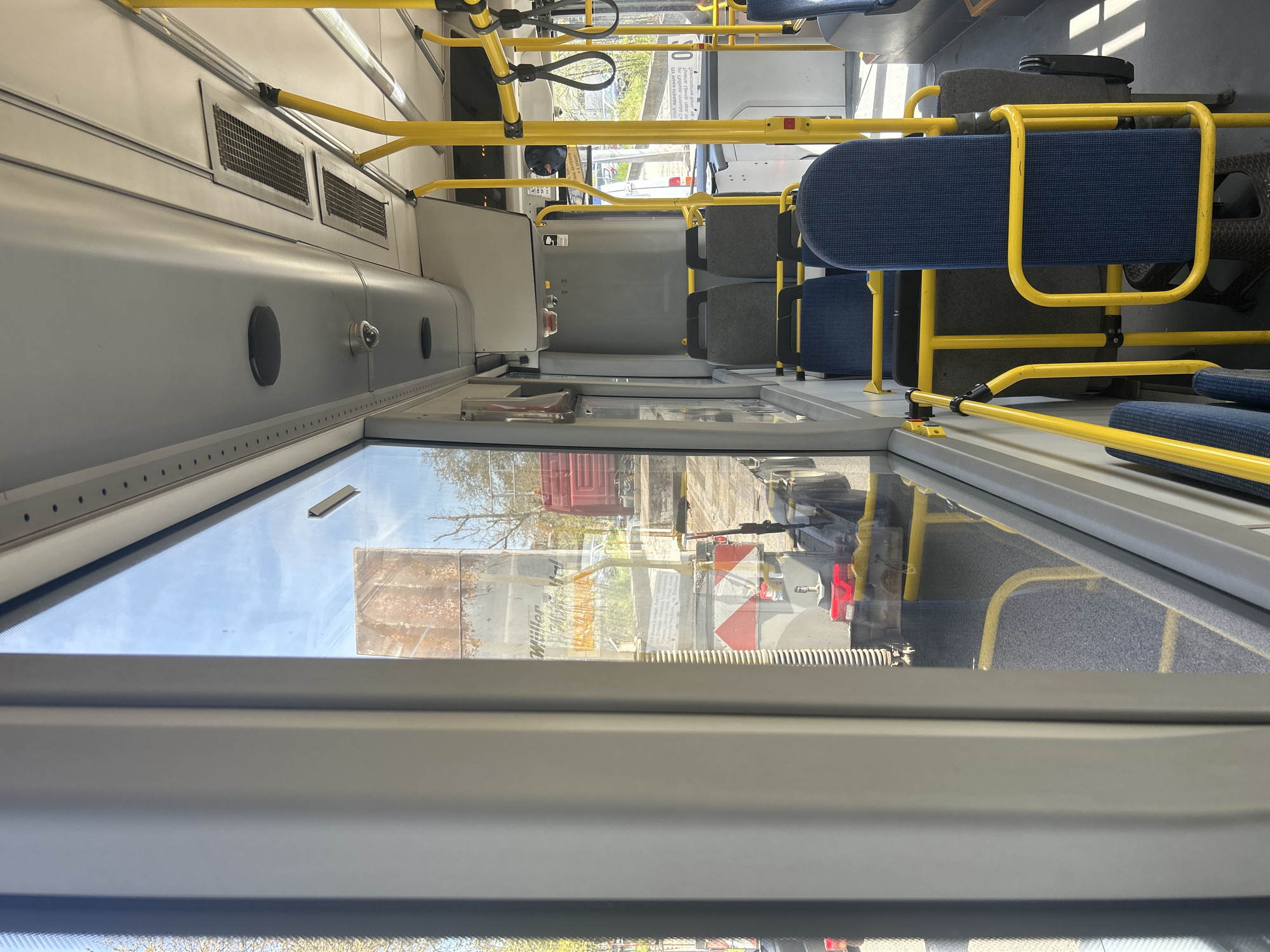 The image depicts the clean interior of a public transport bus, likely in Plovdiv, Bulgaria, viewed from the rear or middle looking towards the front.
On the left side, a large window offers a glimpse of the bright exterior. Through the window, a clear or partly cloudy sky is visible, indicating daytime. Trees with bare branches suggest late winter or early spring. Reflections and direct views outside show parts of several vehicles, including a large red truck and another vehicle with distinctive red and white chevron markings at its rear. The word "Müller" is partially visible in white text on one of the exterior vehicles.
The bus interior features light grey walls and ceiling. Numerous bright yellow grab rails and poles are prominent throughout, installed for passenger safety and support. Some black loop strap handles hang from overhead rails. Standard bus seats are arranged along the right side, upholstered in a mix of dark blue and dark grey patterned fabric. No passengers are visible, suggesting the bus is empty or sparsely occupied.
Notable details on the interior include two rectangular ventilation grilles high on the left wall, along with circular elements that appear to be speakers or lights. A small security camera is mounted on the wall above the seats. Near the front, a white sign with partially legible black text, starting with "UA Home...", is visible, likely providing service information. The floor, visible in parts, appears to be dark grey and textured. The overall bright illumination indicates it is daytime, possibly a sunny day.