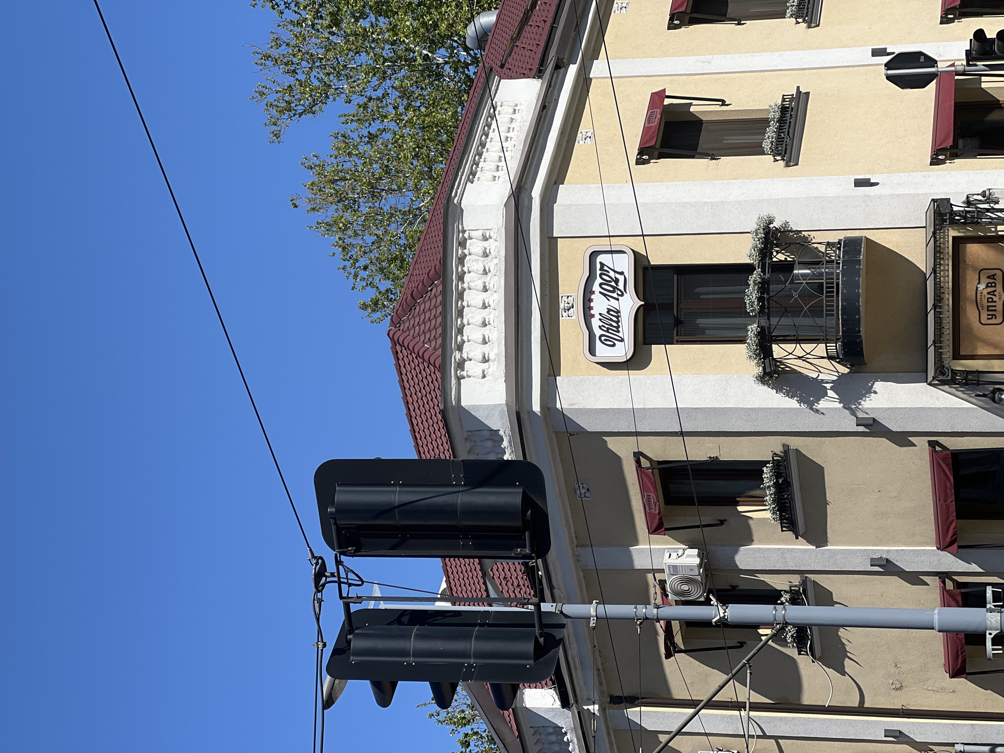 The image shows a street scene in Palilula Urban Municipality, Serbia. The shot is taken from a low angle, looking up at a building and a traffic light. The building has a cream-colored facade with white trim and a red tile roof. There are several windows with dark curtains and flower boxes. A sign reads "Villa 1927" in a stylized font. A gray utility pole with two black traffic lights is in the foreground. A power line stretches diagonally across the blue sky. The lighting suggests it is daytime.