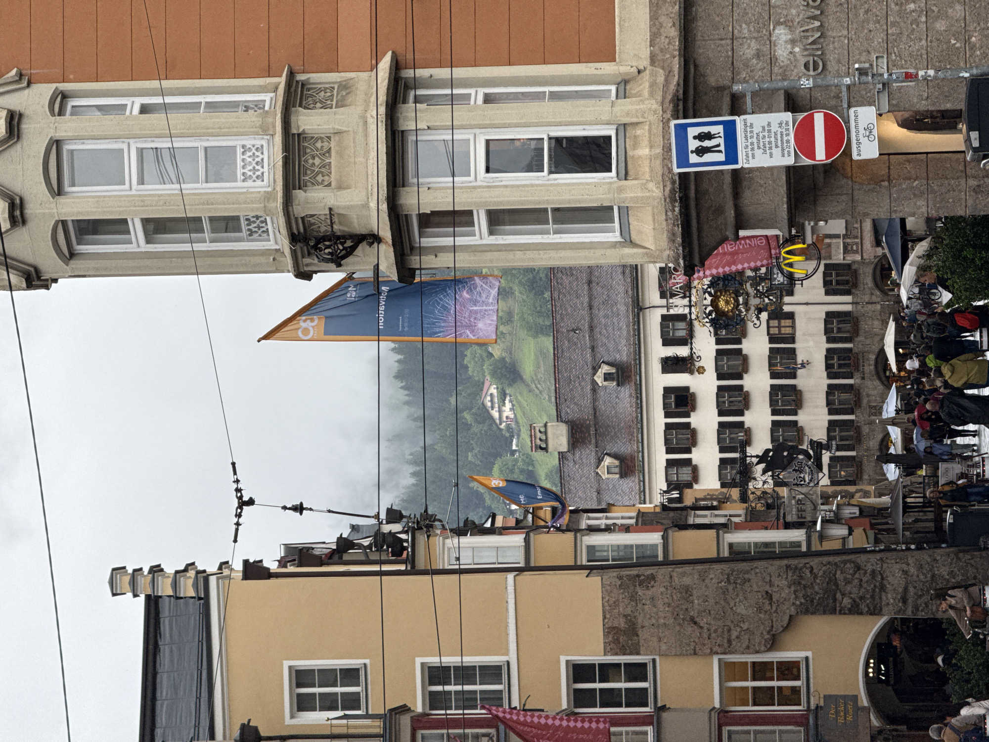 The image captures a street scene in Innsbruck, Austria, on a cloudy day. The architecture features a mix of historical buildings with traditional Austrian facades. On the left, a yellow building with a distinct stepped gable and white-framed windows dominates the foreground. To the right, a multi-story building with an orange facade and a cream-colored bay window is visible. A large blue banner with fireworks imagery hangs from this building.
Below these buildings, a bustling street with pedestrians is depicted. People are seen walking, some carrying umbrellas, suggesting intermittent rain. Several shops and businesses line the street, indicated by various signs, including a McDonald's logo and a sign with German text reading "Der Bäcker Ruetz." Street lamps and overhead wires are prominent features of the urban environment. In the background, a lush green hillside is dotted with houses and trees, partially obscured by mist, indicating the city's proximity to the Alps. Traffic signs are also visible, including a sign for restricted access and a red circular prohibitory sign.