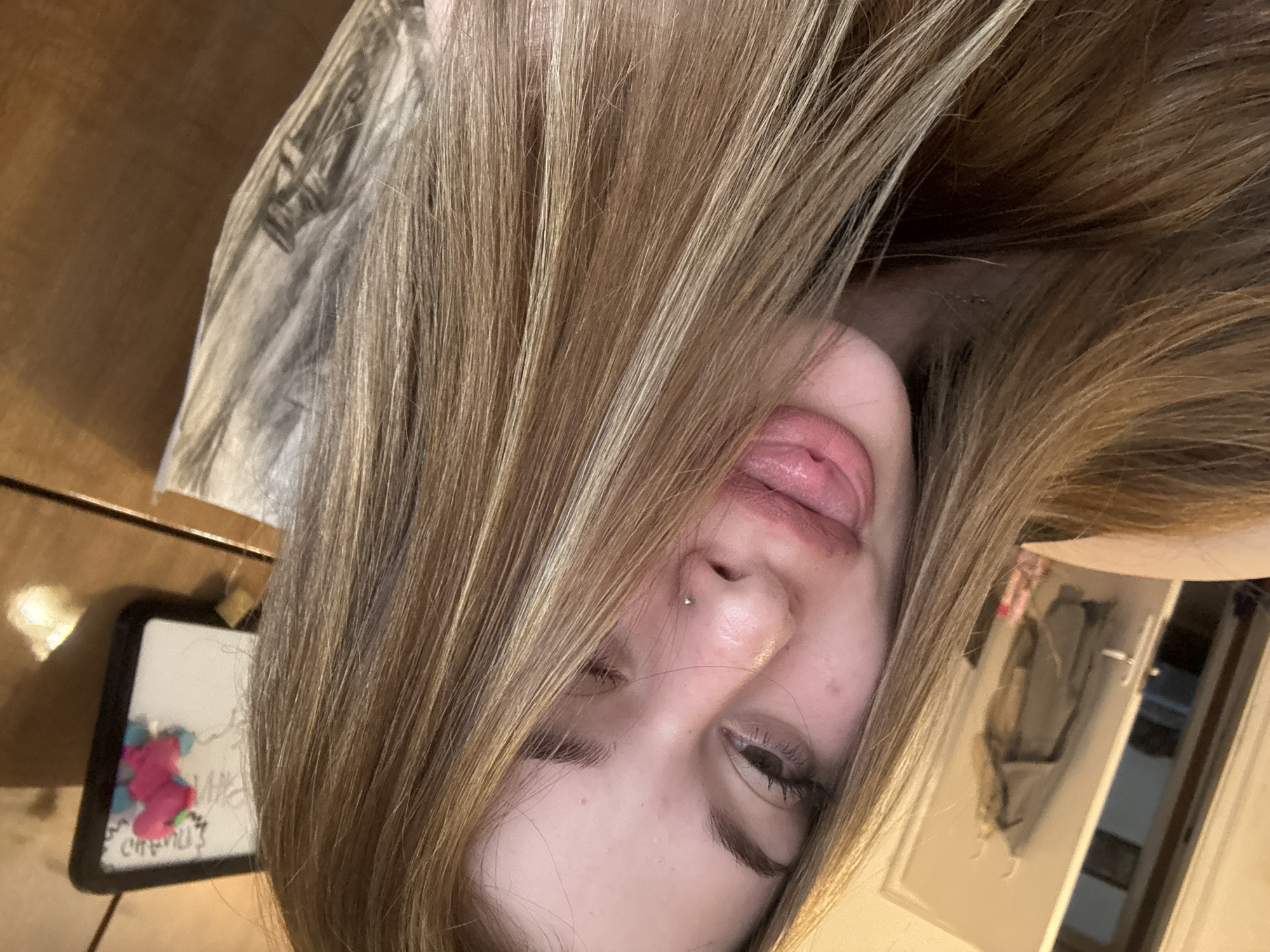 A close-up selfie, taken indoors with the subject's face predominantly in the frame, is angled upside down. The subject is a young individual with light brown hair, partially obscuring the face. The subject's tongue is slightly extended. A nose piercing is visible on the subject's left nostril. One eye is partially closed in a wink.
In the background, there is a wooden surface, possibly a table, with a small, framed object on it. Further back, a white cabinet is visible. The location, Troyan, Bulgaria, is not discernable from the visual information provided.