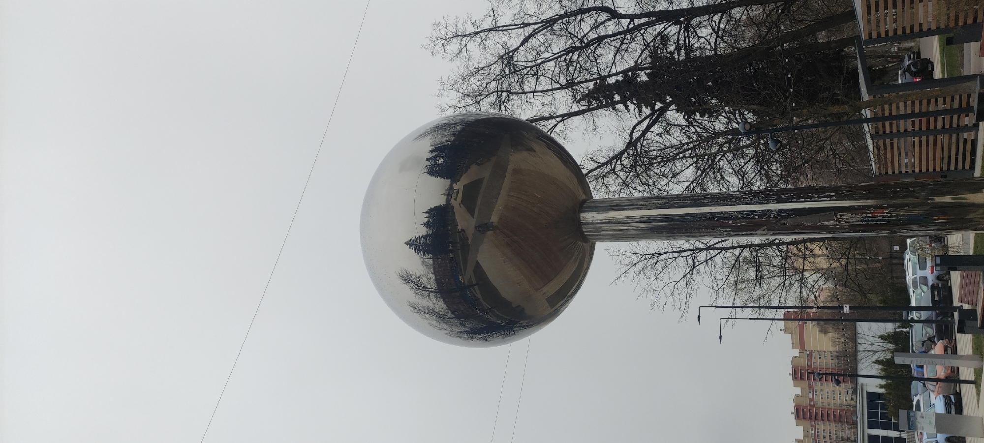 The image shows a large, highly reflective metallic sphere mounted on a post, likely a public art installation. The sphere reflects the surrounding environment. In the reflection, one can discern trees, buildings, and the general landscape. The sky is overcast. Bare trees are visible in the background above the sphere, and a building and parked cars are seen on the ground level. Based on visual clues and cross-referencing with online resources, this art installation is likely located in Fryazino, Russia. The surrounding elements include city architecture, utility poles, and possibly street furniture (benches, etc.). There's no specific indication of human presence near the sphere.