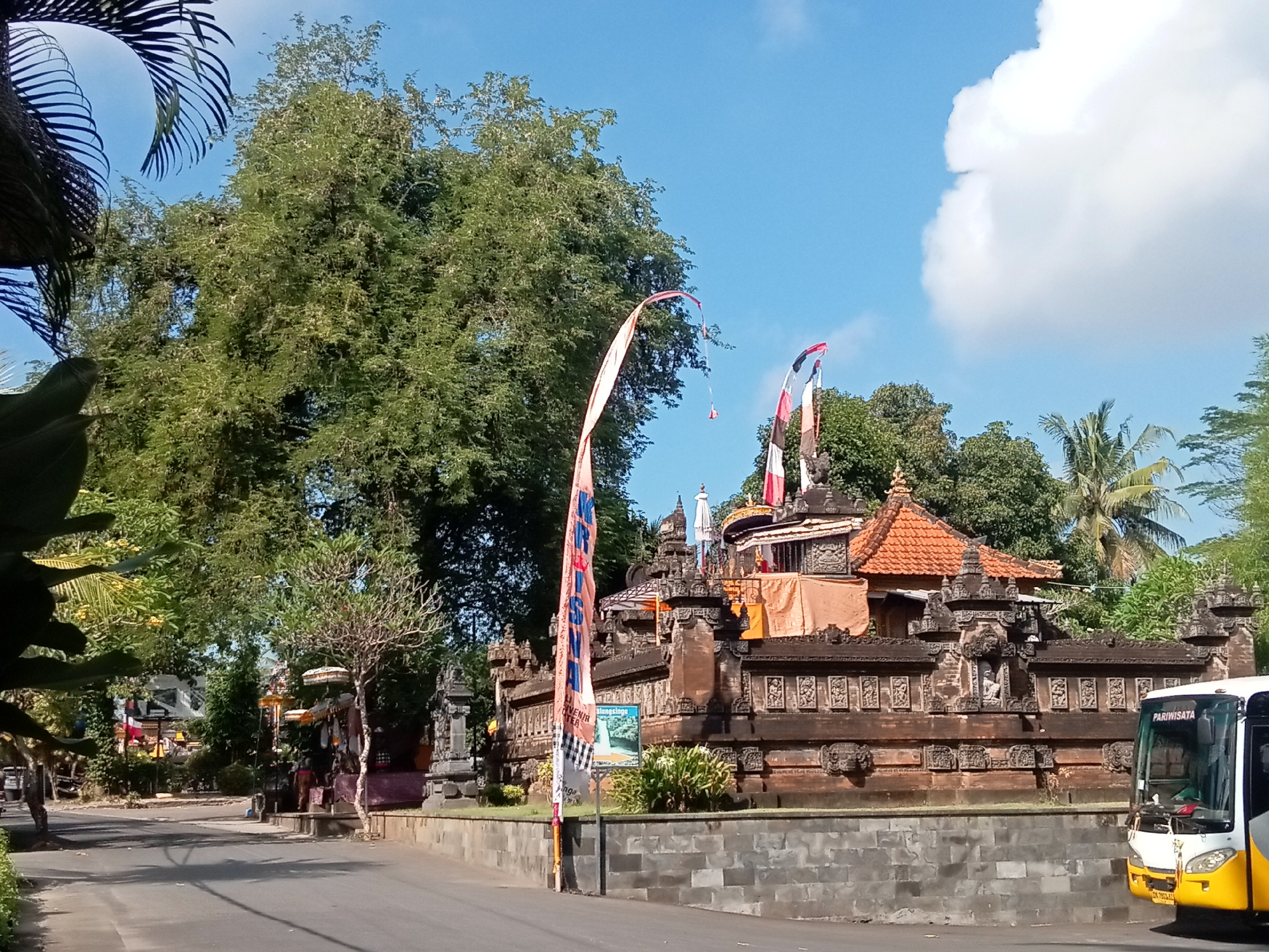 In an outdoor daytime scene, a bus with the text "PARIWISATA" on its windshield is parked on the right side of a paved road. To the left of the road is a stone wall, followed by a building with traditional Balinese architecture, featuring intricate carvings and an orange tiled roof. Several decorative flags with red and white patterns are erected in front of the building. Tall green trees with dense foliage are visible in the background, and palm fronds can be seen on the left edge of the image. The sky is blue with some white clouds. The scene appears to be a cultural site or attraction in a tropical or subtropical location.