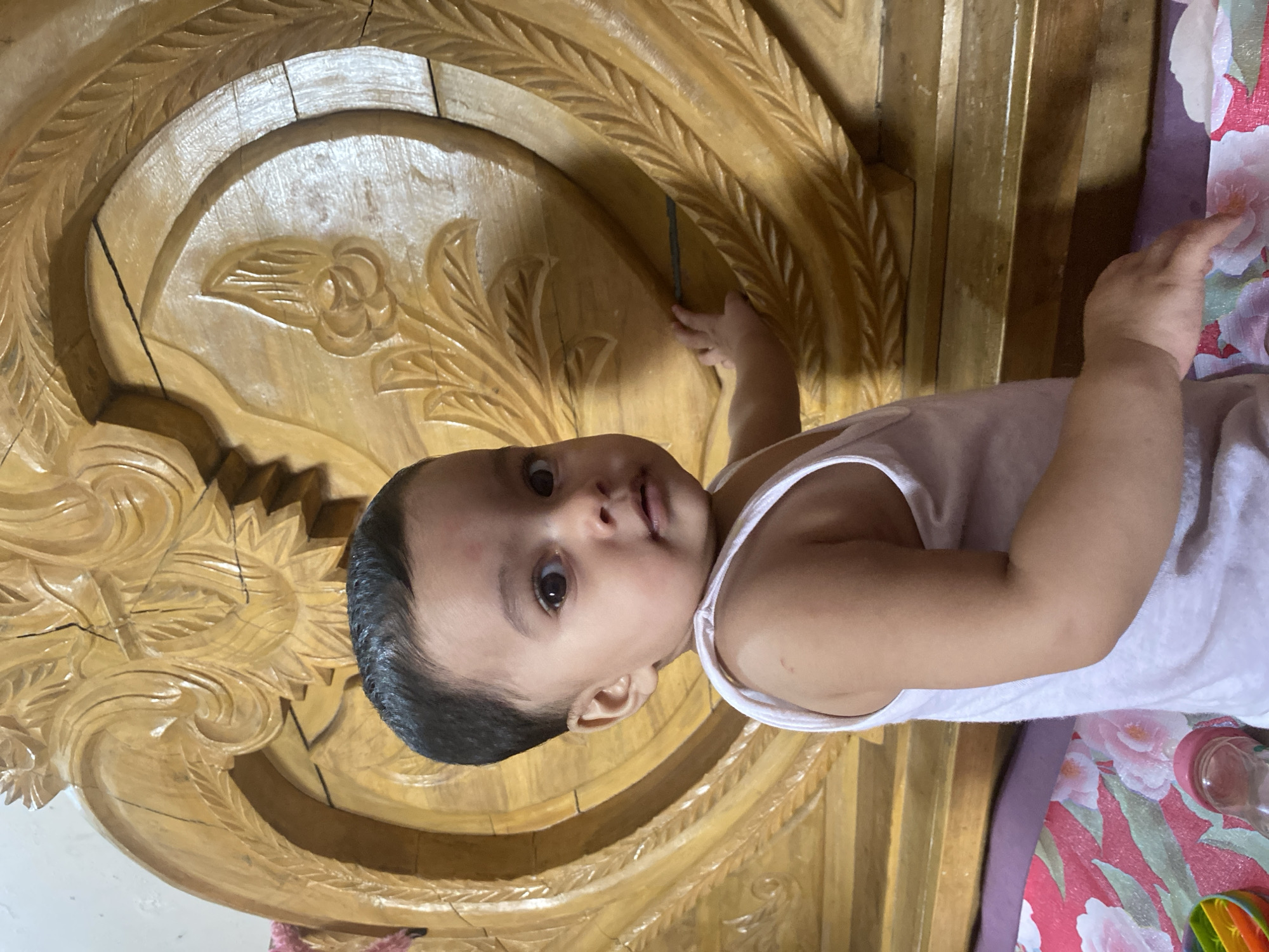 A close-up, eye-level portrait shows a baby, likely an infant or toddler, with dark hair and brown eyes, looking directly at the camera with a neutral to slightly curious expression. The baby wears a simple white sleeveless top. The scene is indoors, with the baby positioned next to a substantial piece of light-colored, ornately carved wooden furniture, which appears to be a headboard or a decorative wall panel. The wood features intricate floral and leaf carvings. The baby's left hand is visible, reaching towards the carved wooden surface. To the right of the baby, a floral patterned fabric in shades of pink, white, and red/purple is partially visible, along with a clear baby bottle or cup and a small portion of a colorful toy. The setting is consistent with a home interior in Dhaka, Bangladesh. No text is visible in the image.