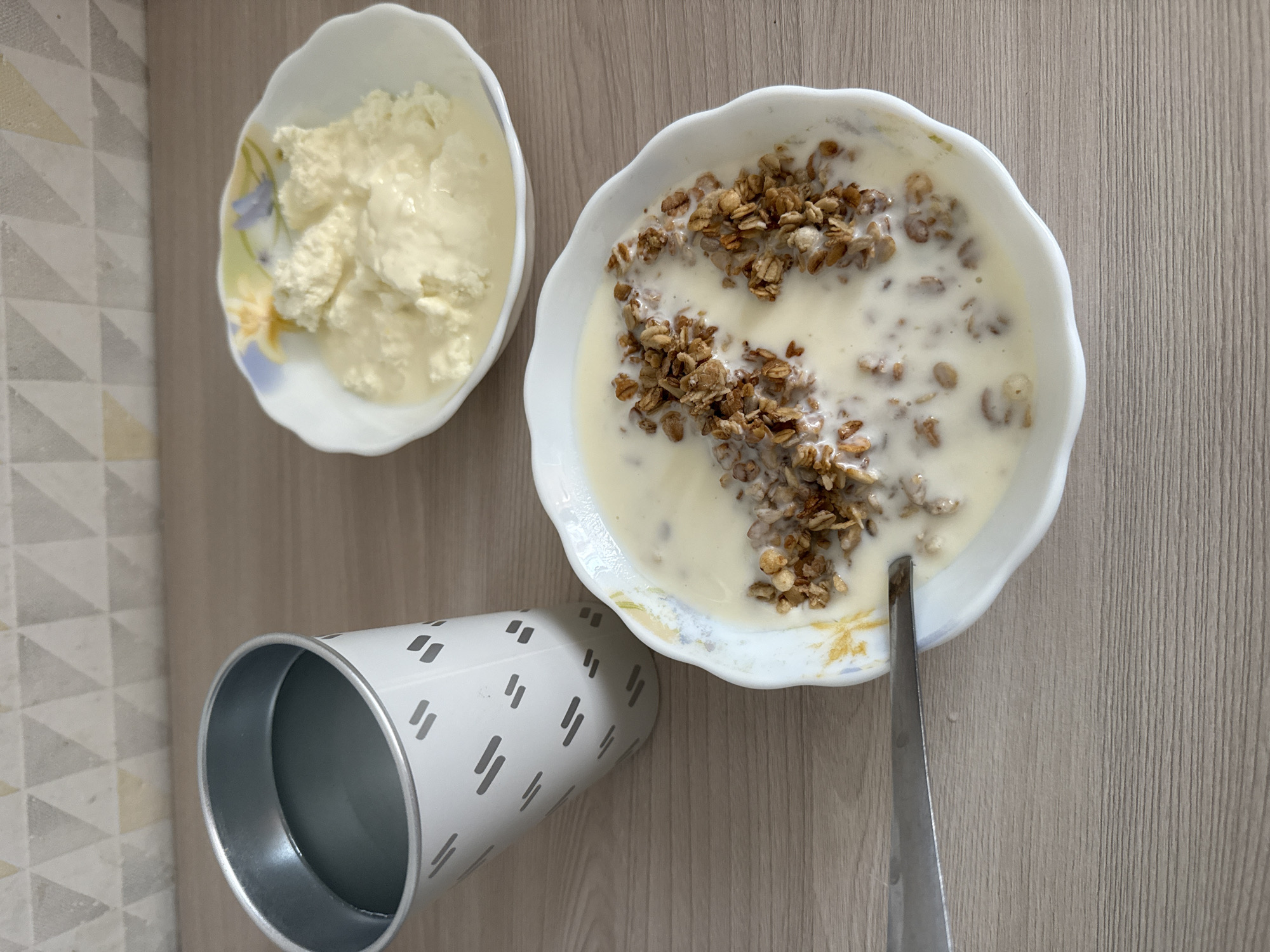 On a light brown wooden surface, a meal is laid out. In the foreground, a white bowl with a scalloped rim contains a creamy white substance with scattered granola clusters. A silver spoon rests on the edge of this bowl, with its tip submerged in the food. Behind this, to the right, is a smaller white bowl containing a dollop of cottage cheese or ricotta, swimming in a light liquid. To the left, a white cylindrical cup with a silver rim features a pattern of diagonal gray dashes. The cup is tilted and contains a dark liquid. The background shows a wall with a geometric pattern of light brown, cream, and beige triangles. The lighting suggests an indoor setting.