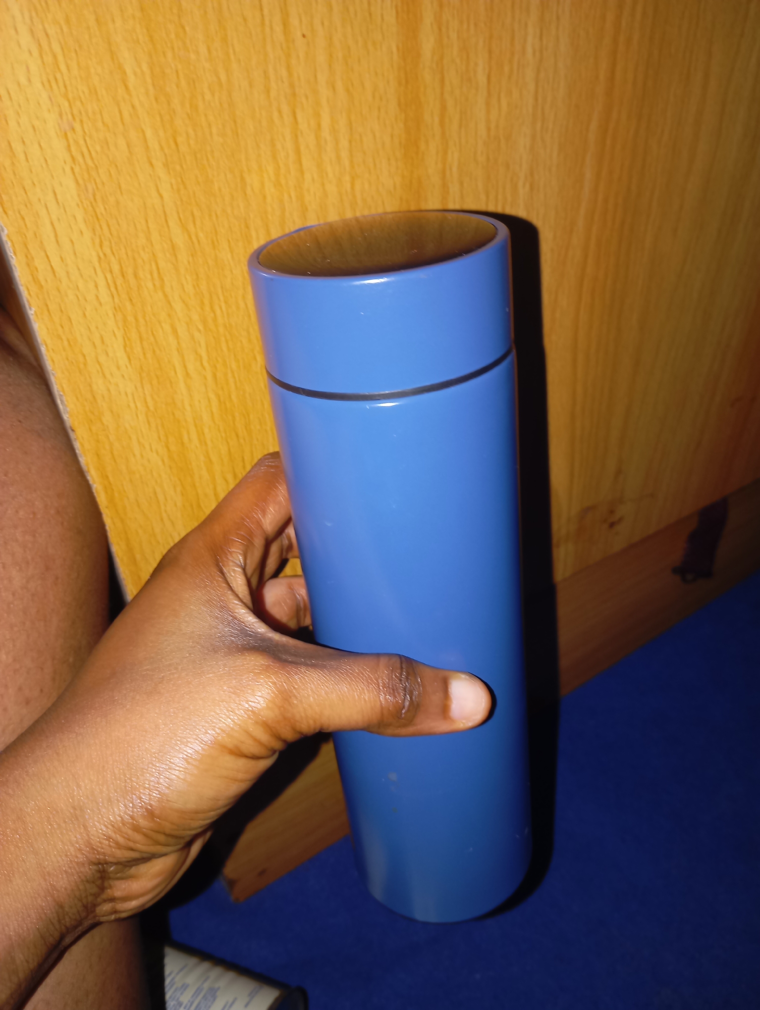 A hand holds a blue cylindrical thermos against a wooden background. The thermos is upright and appears to be made of metal. The hand is dark-skinned and is gripping the thermos around its middle. The background is a wooden surface with a visible grain pattern. The lighting suggests an indoor environment.
