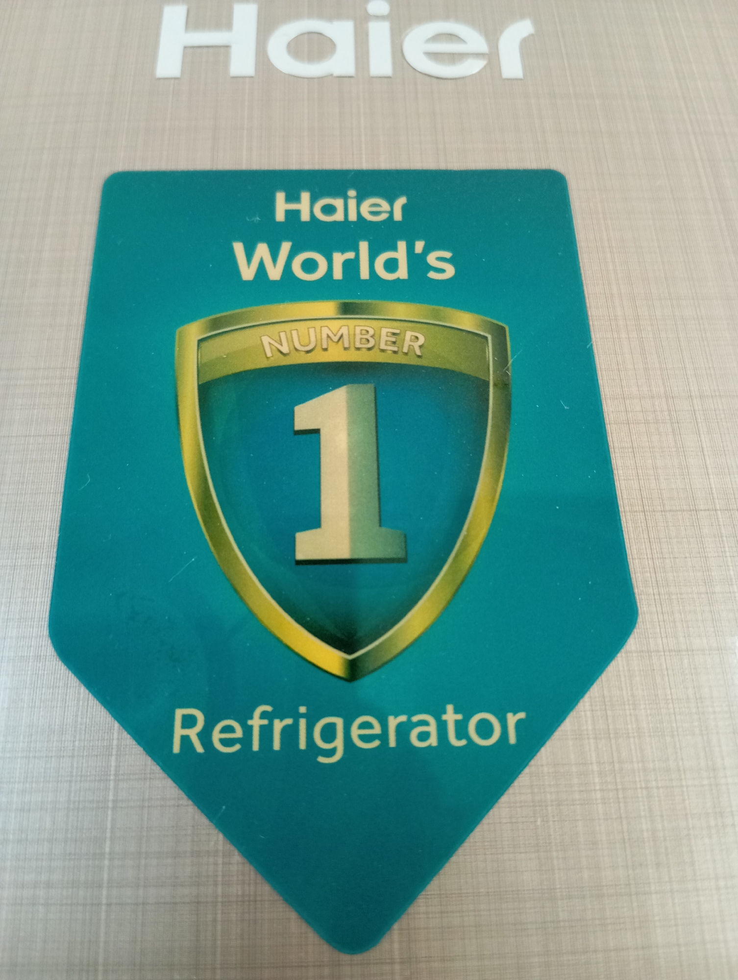 A close-up, high-angle, full shot shows a teal-colored shield-shaped sticker with gold trim on a light gray, textured surface. At the top of the sticker, the word "Haier" is written in large, white, sans-serif capital letters. Below this, also in white text, is "Haier World's NUMBER 1 Refrigerator." The number "1" is prominently displayed in large, light yellow, block-style font within the shield. The textured surface appears to be a metal appliance.