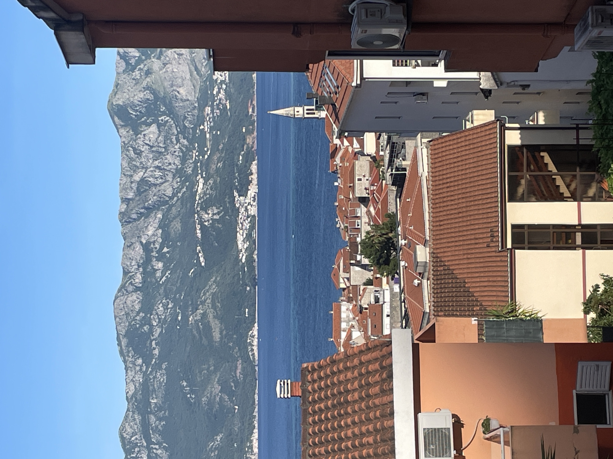 The image depicts a view in Budva, Montenegro, with a landscape that includes mountains, the Adriatic Sea, and the red-tiled rooftops of the city. A notable tower is visible among the buildings. Buildings and other rooftops are in the foreground. The sky is clear and blue.