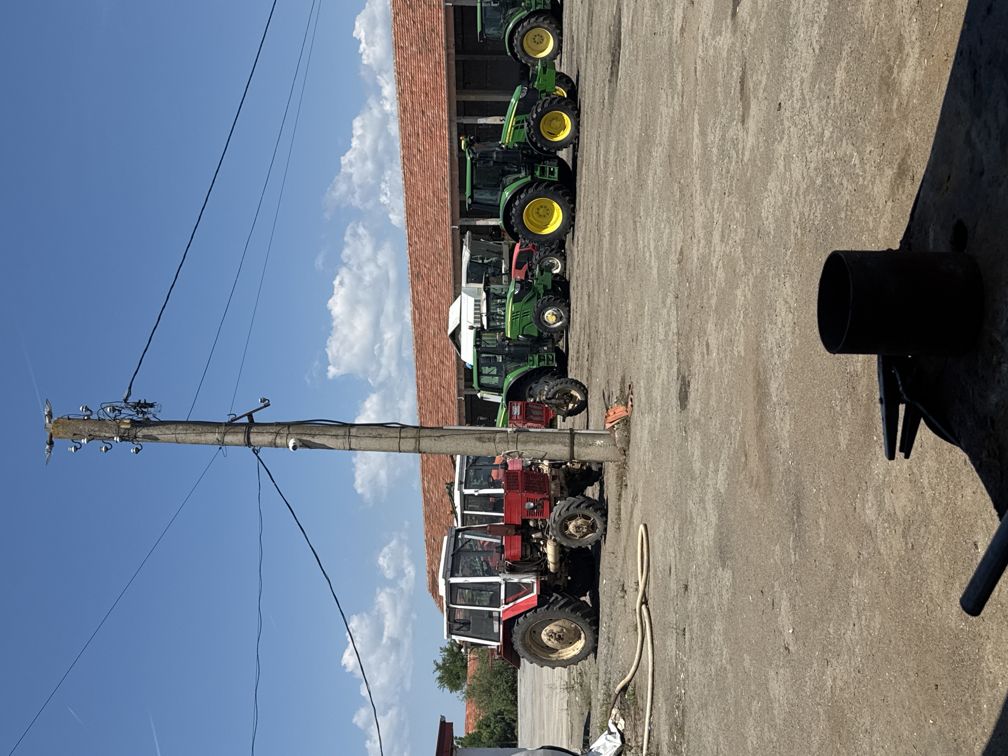 The image shows a row of parked tractors in Sarnevo, Bulgaria, along a concrete yard in front of a building with a red tiled roof. Tractors of varying colors and models are parked in a row. A concrete utility pole stands in the foreground, with electrical lines extending across the blue, partly cloudy sky. A section of black pipe, possibly part of a vehicle, is visible on the concrete in the foreground. No people are visible in the image.