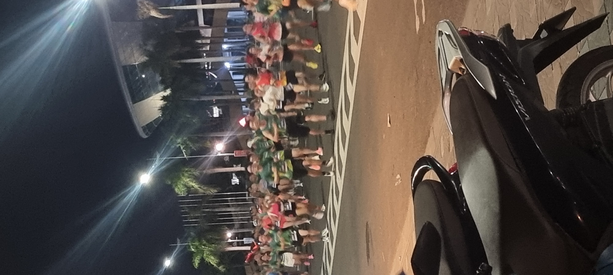 A large group of runners are participating in a nighttime race on a paved road. The runners are wearing athletic clothing, predominantly green and white, and many have bib numbers. Palm trees and streetlights line the road, with a modern building, possibly a stadium or convention center, visible in the background under a dark sky illuminated by artificial lights. In the foreground, a black motorcycle is parked on the side of the road. The image appears to be taken in Đà Nẵng, Vietnam, as suggested by the presence of palm trees and the architectural style of the building.