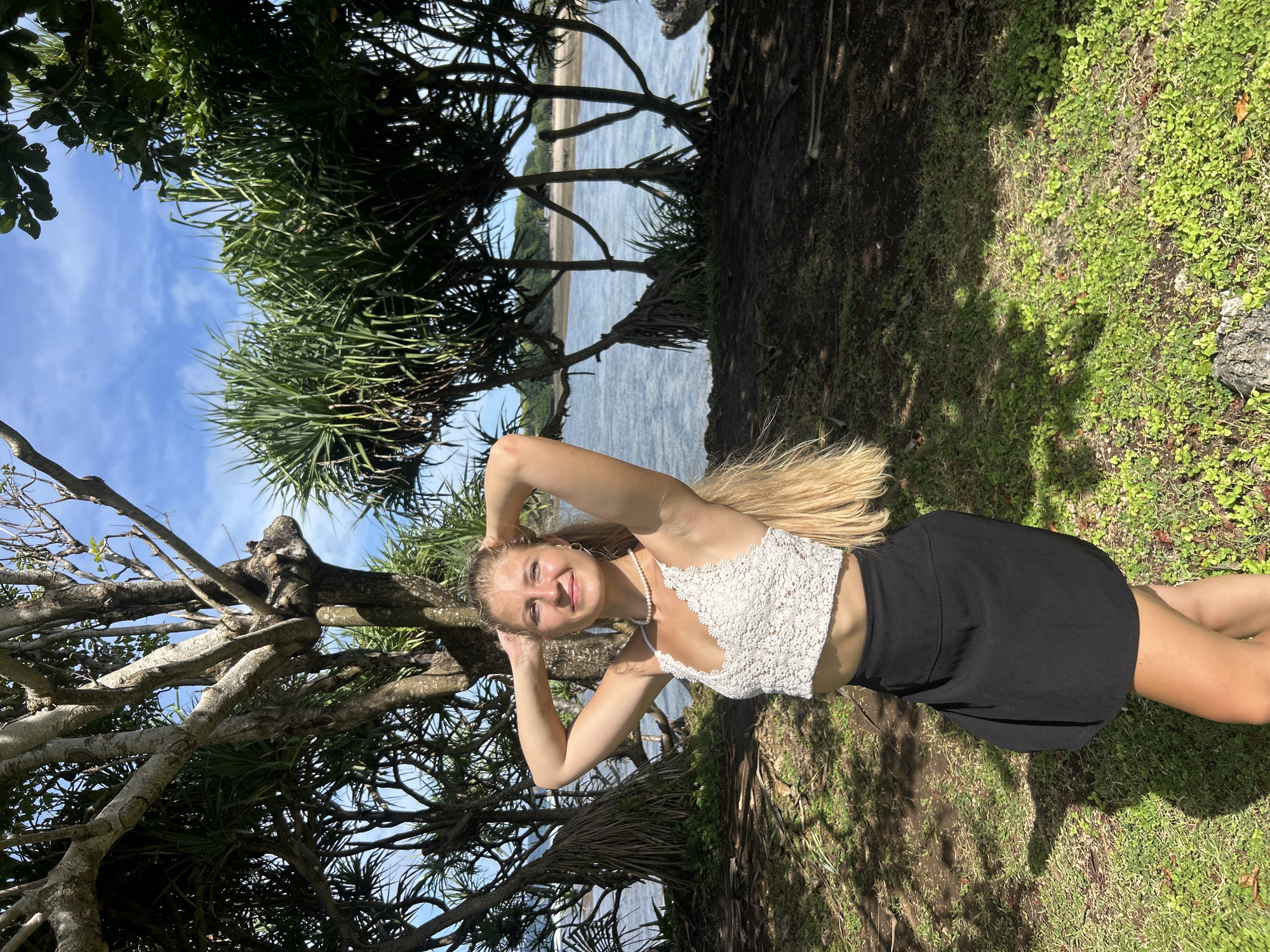 A young woman with long, blonde hair stands outdoors, smiling directly at the camera with her hands clasped behind her head. She is wearing a white crocheted or lace crop top, a pearl necklace, and a black mini skirt. Her expression is joyful and relaxed.
The setting appears to be a tropical coastal area. She is positioned on a grassy patch with visible dirt and shadows from the surrounding trees. Behind her, dense, spiky-leaved tropical trees, likely Pandanus, with gnarled branches, frame the scene. These trees extend into the background, where a body of water—possibly the ocean or a bay—can be seen, with a sandy shoreline and distant land visible beyond. The sky above is clear and blue with a few scattered white clouds.
The lighting indicates a bright, sunny day, likely mid-morning or afternoon, casting distinct shadows on the ground. The overall atmosphere is vibrant and natural. The tropical vegetation and coastal landscape are consistent with the provided location context of Kuta Selatan, Indonesia. There is no visible text in the image.