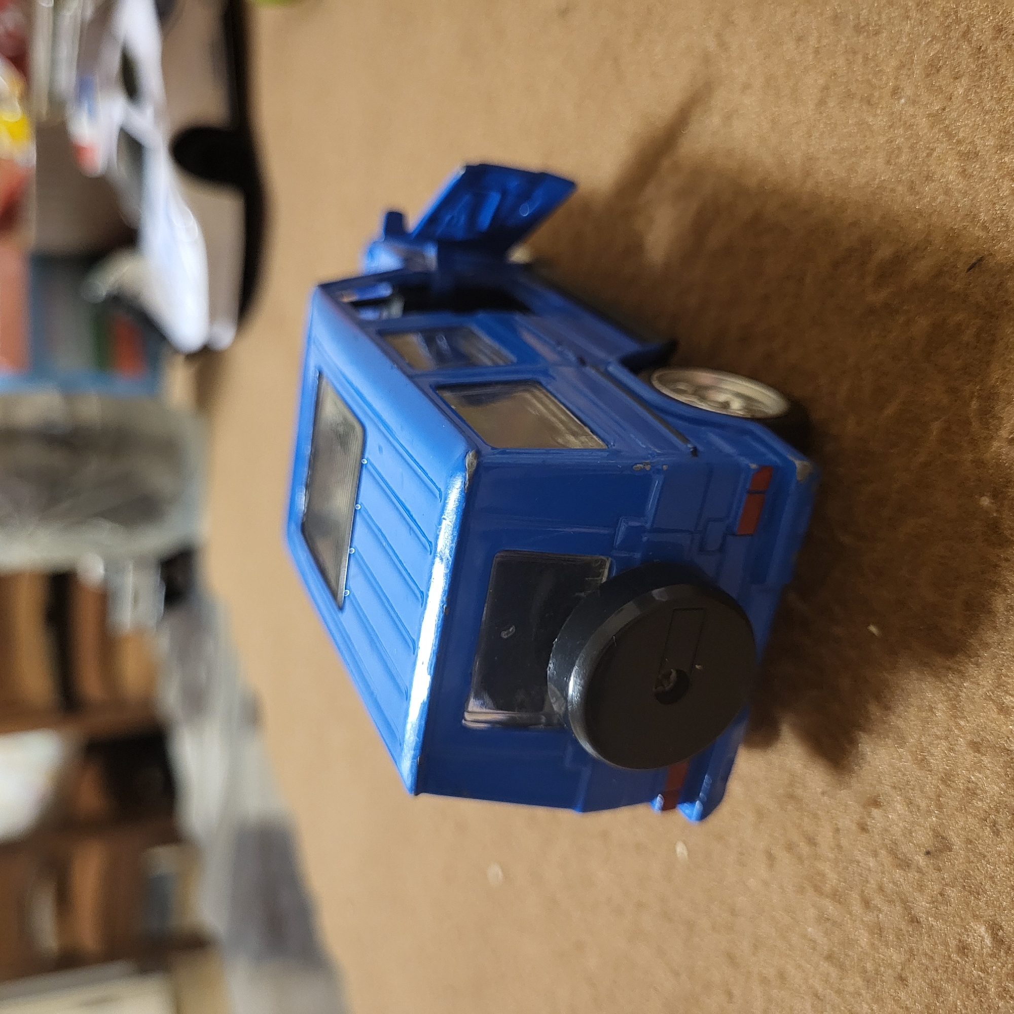 A blue toy SUV is situated on a beige surface, seemingly indoors. The toy's hood is open. Other objects, including what appears to be a toy car, are visible in the blurry background. The location is not discernible from the image.