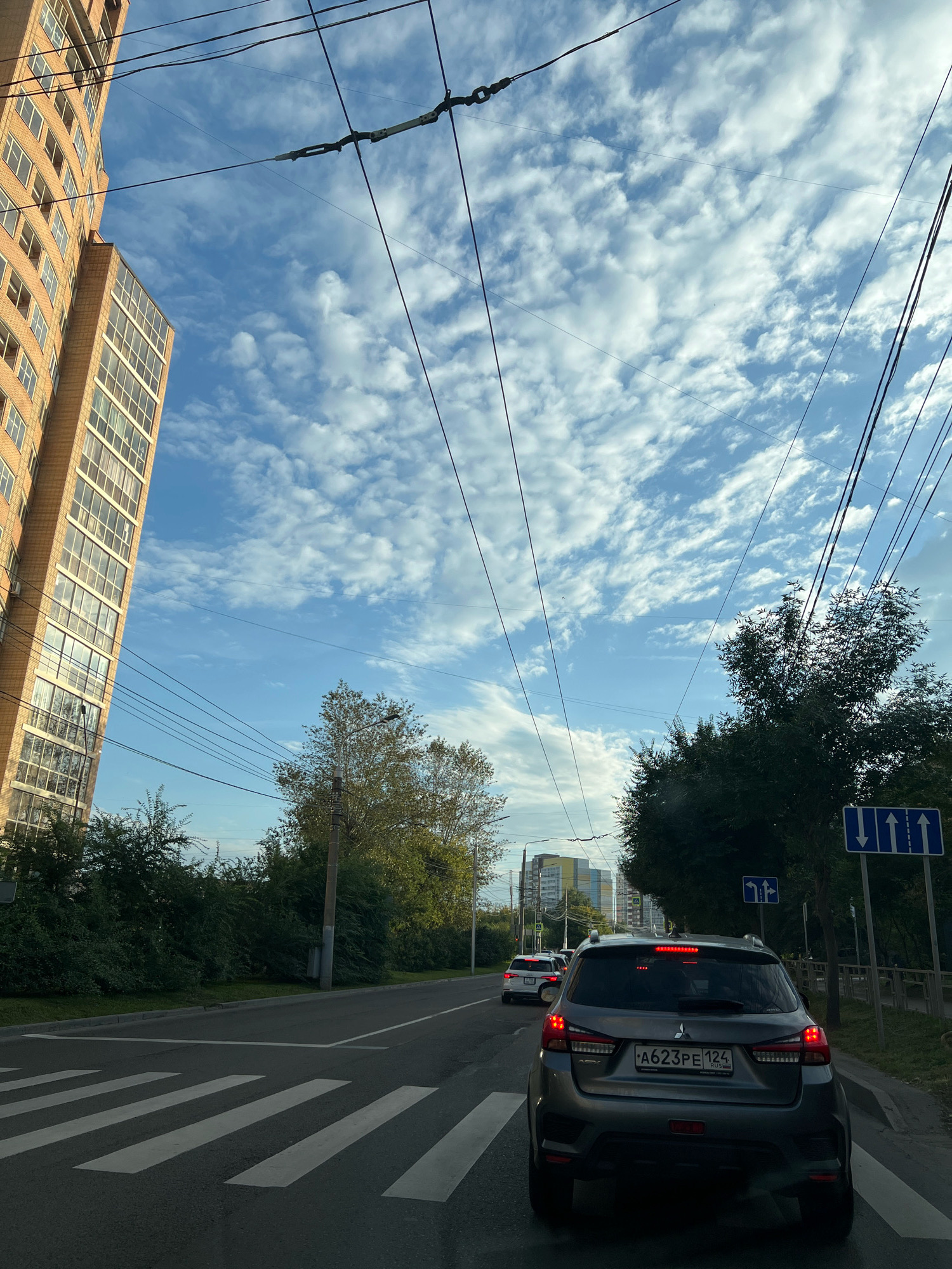 The image shows a view from inside a car driving on a street in Krasnoyarsk, Russia. The car is stopped at a pedestrian crossing. In the foreground is the rear of a grey Mitsubishi crossover SUV, license plate A623PE124. The car has its brake lights on. Further ahead, other vehicles are visible in traffic. To the left, a tall, multi-story apartment building with a brick facade and numerous windows is prominent. The sky above is blue with scattered white clouds. Overhead, a network of power lines or trolley wires crisscrosses the sky. Trees line the right side of the road, and a street sign with three upward arrows is visible. The overall scene depicts a typical urban streetscape during daylight hours.