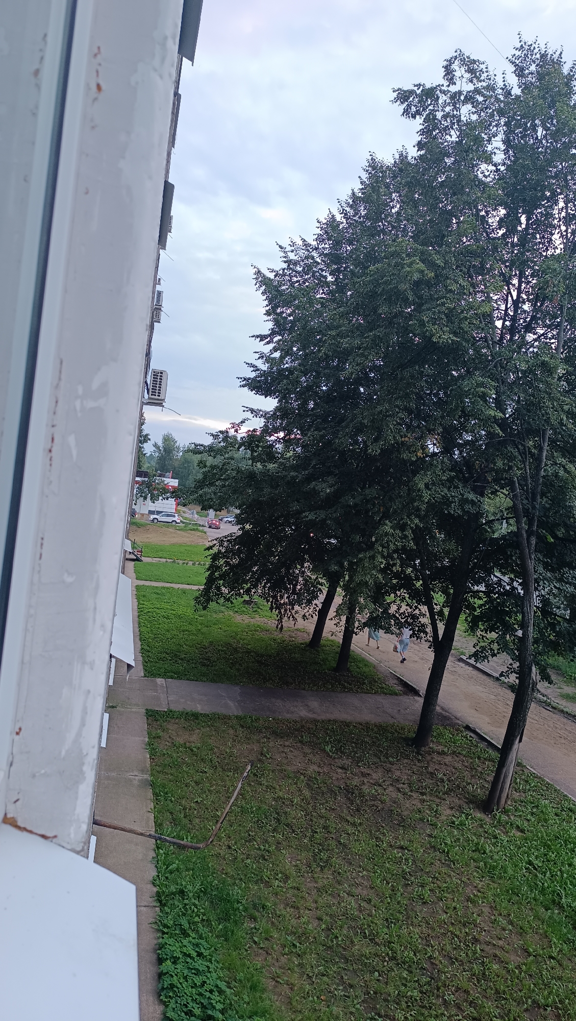 This is an outdoor, eye-level, medium shot photograph taken from a building's window in Chaikovsky, Russia. The left side of the frame is dominated by the exterior wall of the building, painted white with some peeling and discoloration. Visible on the wall are several air conditioning units. In the foreground, a metal pipe protrudes horizontally from the building wall and angles downwards. Beyond the building's immediate vicinity, a patch of mowed grass transitions to a dirt pathway. Several trees with dense green foliage are scattered across the midground and background. In the distance, cars are parked along a road, and two people are walking along the pathway under the shade of the trees. The sky above is overcast with diffused light, indicating either early morning or late evening.
