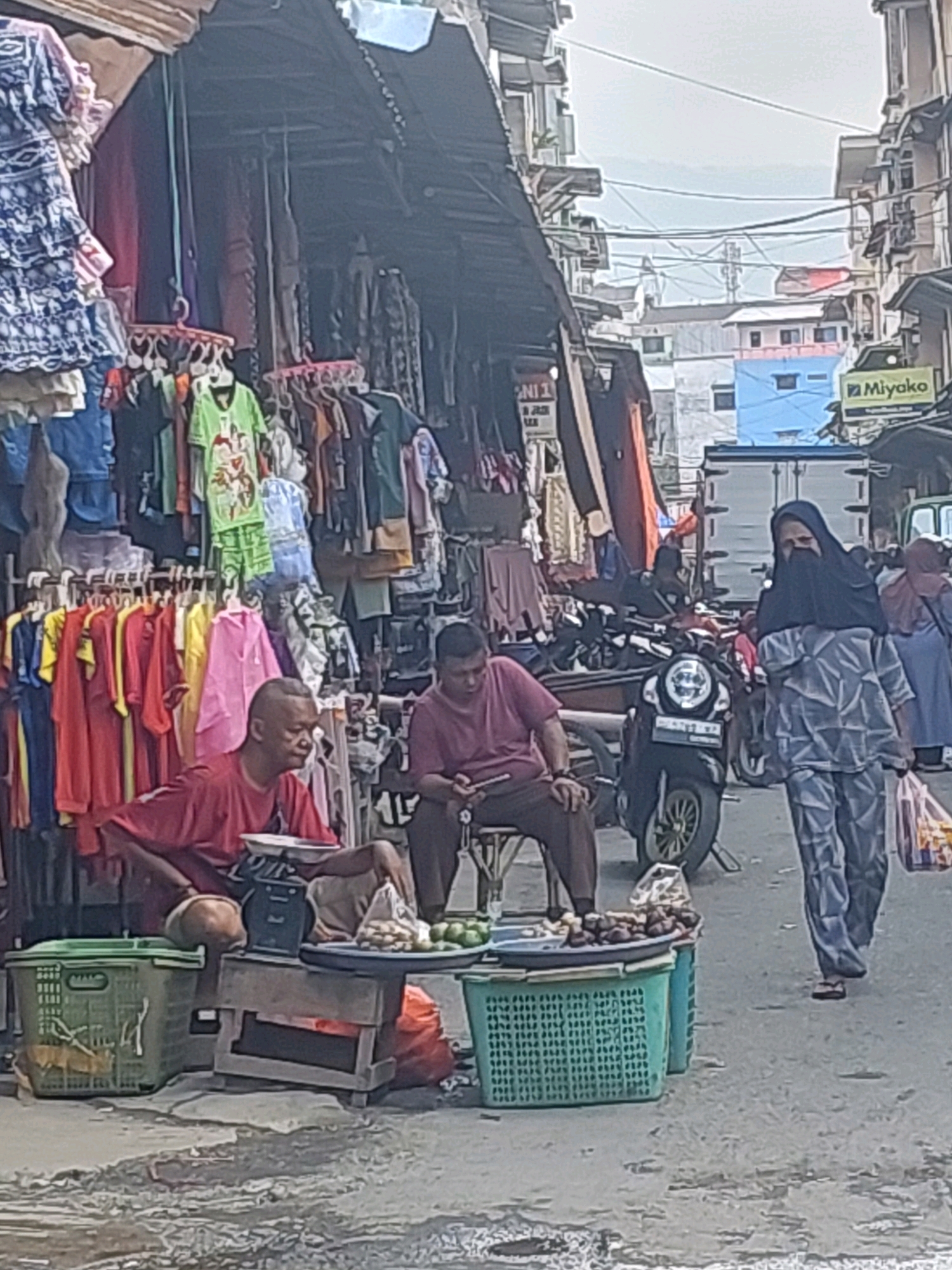 The image captures a street scene in Ambon, Indonesia, likely a marketplace. In the foreground, two men are seated at a stall laden with goods. One man, wearing a red t-shirt, is leaning forward, looking to his right. Beside him, a weighing scale is visible. The other man, in a maroon t-shirt, is looking down at his phone. A variety of items, including fruits and what appear to be nuts or small vegetables, are displayed on platters resting on green plastic baskets. Behind them, a stall displays an assortment of clothing items hanging on racks. In the background, a woman wearing a black hijab and patterned gray clothing is walking away from the viewer, carrying a plastic bag. A motorcycle is parked near the stall. Further in the background, buildings and a truck are visible, suggesting an urban environment. The ground is paved and appears wet in places, possibly from recent rain. The overall atmosphere suggests a busy, everyday market setting.