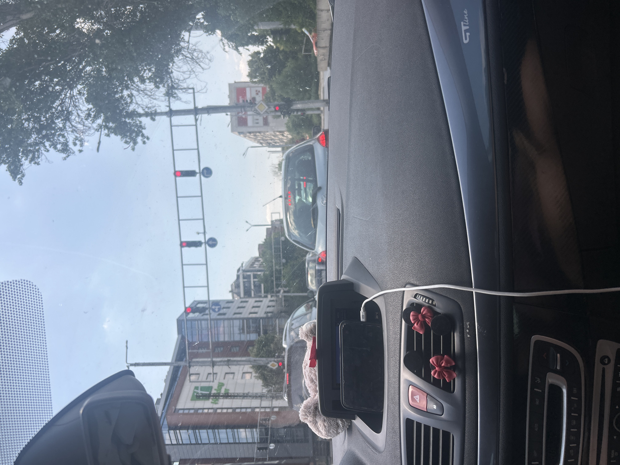 The image is taken from inside a car, looking out through the windshield, on a bright, cloudy day. The car is stopped at a traffic light in what appears to be an urban environment. Several traffic lights are visible, some showing red. Buildings of various heights and architectural styles line the street, including a modern building with a "Holiday Inn" sign. Trees are present on the right side of the frame. The car's dashboard is visible in the foreground, with a phone charging and a fluffy toy with red bows attached to the air vents. The car is a Renault, as indicated by the "GT Line" badge visible on the interior trim. The license plate of the car in front is not visible. The specific location in Plovdiv, Bulgaria cannot be determined from the image alone.