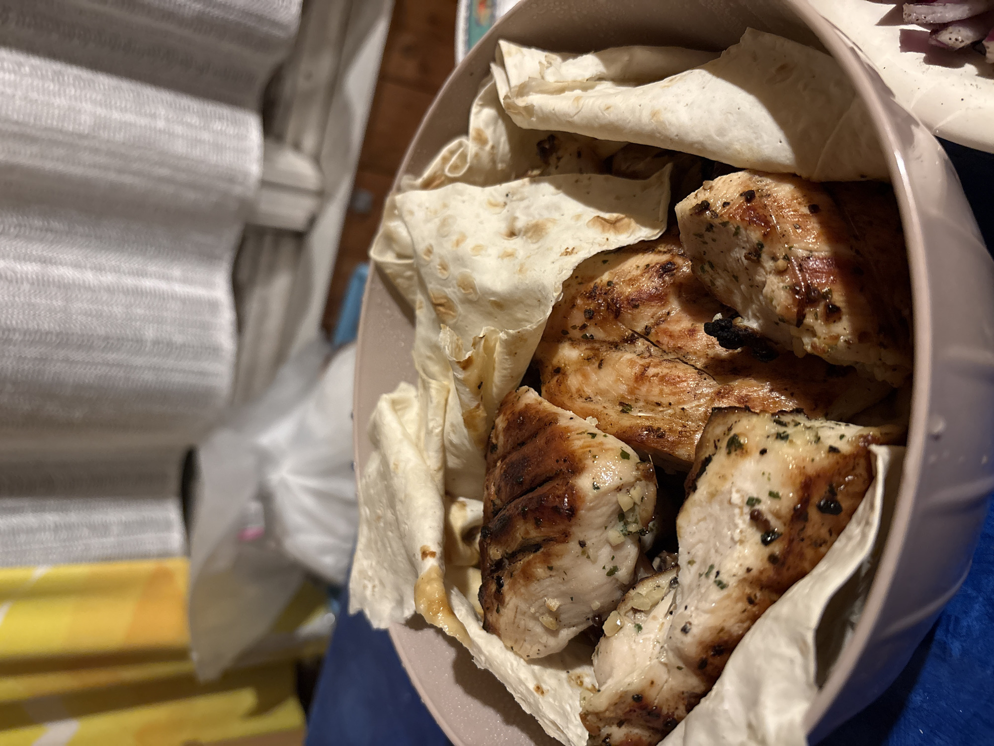 A close-up, eye-level shot shows grilled chicken pieces nestled within pieces of pita bread in a pink bowl. The chicken is seasoned with herbs and has grill marks. The background is blurred, with hints of yellow and white curtains. The scene is indoors. The image does not depict a location in Пярэжырскі сельскі Савет, Belarus.