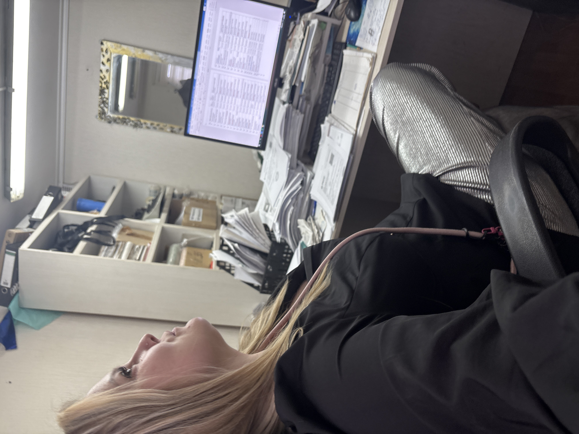 A woman is lying down in an office setting. She has blonde hair and is wearing a black top. Her legs, clad in silver leggings, are visible next to her. The office has a computer monitor displaying text, shelves filled with files and boxes, and a desk covered with papers. A mirror is on the wall. The location is Noviy, Russia.