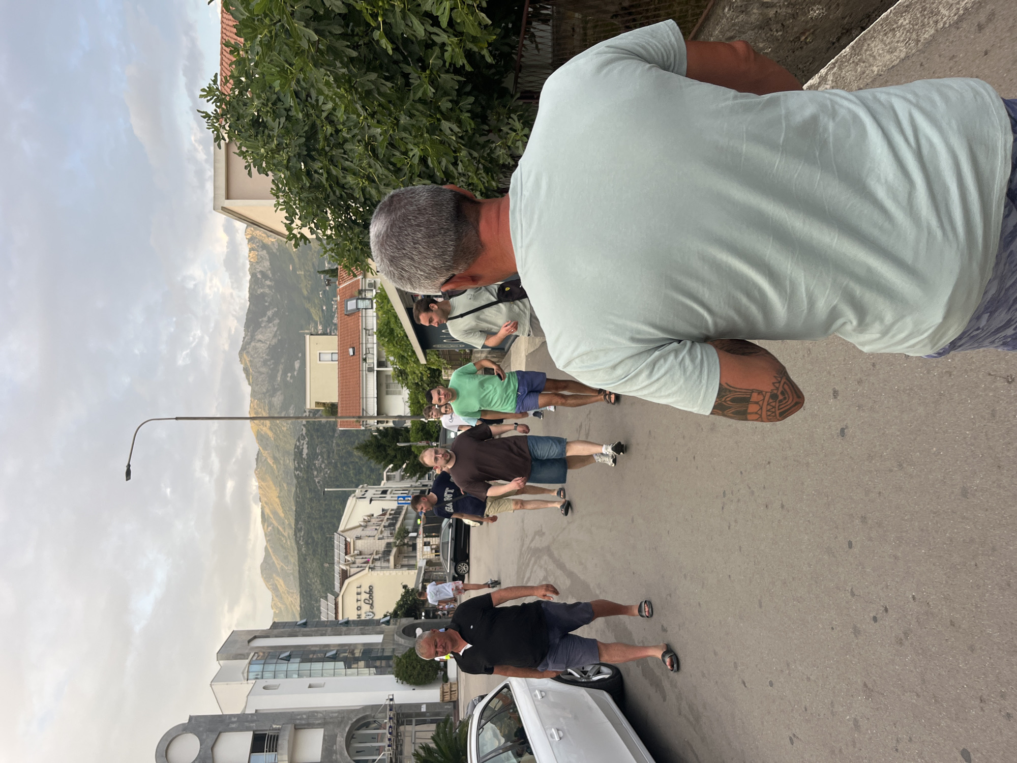 In this eye-level medium shot, a group of men are walking down a paved street in what appears to be a town or city. The perspective is from behind one of the men, who is facing away from the camera. He has short, gray hair and is wearing a light blue t-shirt and patterned shorts. A tattoo is visible on his left arm. The street is lined with buildings on either side, some of which appear to be hotels or businesses, with a mountain range visible in the background. The sky is cloudy and appears to be either late afternoon or early morning. The man in the foreground is walking towards the viewer's right, with other men in the group walking ahead of him and to his left. One man in the foreground, on the left side of the street, is wearing a black polo shirt and shorts. He is walking in the same direction as the man in the foreground. A white car is parked on the left side of the street.