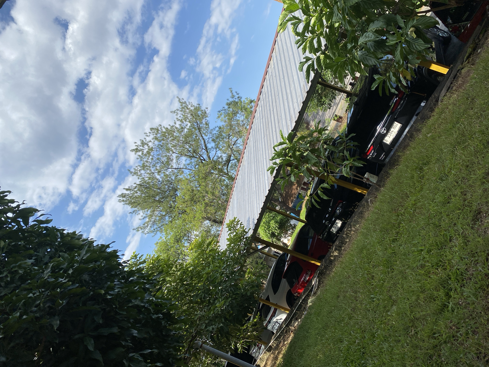 A low-angle shot shows a covered parking area with several cars parked underneath a corrugated metal roof. The cars visible are a white SUV, a red hatchback, and at least two black sedans. The parking area is bordered by a grassy slope in the foreground and trees and sky in the background. The sky is blue with scattered white clouds. The scene appears to be outdoors, possibly in a residential or commercial area. The license plates are partially visible on the black sedans, one reading "B 1564 HOZ". The overall impression is of a sunny day with good visibility. The location is identified as Banjarbaru, Indonesia.
