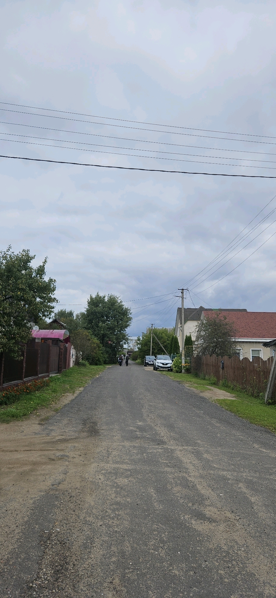 The image depicts a street in a rural or suburban area, possibly in Vitsebsk, Belarus, under an overcast sky. The road is unpaved, composed of gravel and dirt, with some visible patches and wear. On the left side of the road, a brown wooden fence with some greenery and flowers borders a property. Further down the road on the left, there are several trees. On the right side, a wooden fence lines the properties, with houses and vegetation visible behind it. A house with a red roof is prominent on the right. Several cars are parked along the right side of the road, including a white Renault. Two individuals are walking away from the camera down the center of the road, carrying backpacks. Overhead, power lines crisscross the sky. The overall scene suggests a quiet, everyday setting in a settlement.
