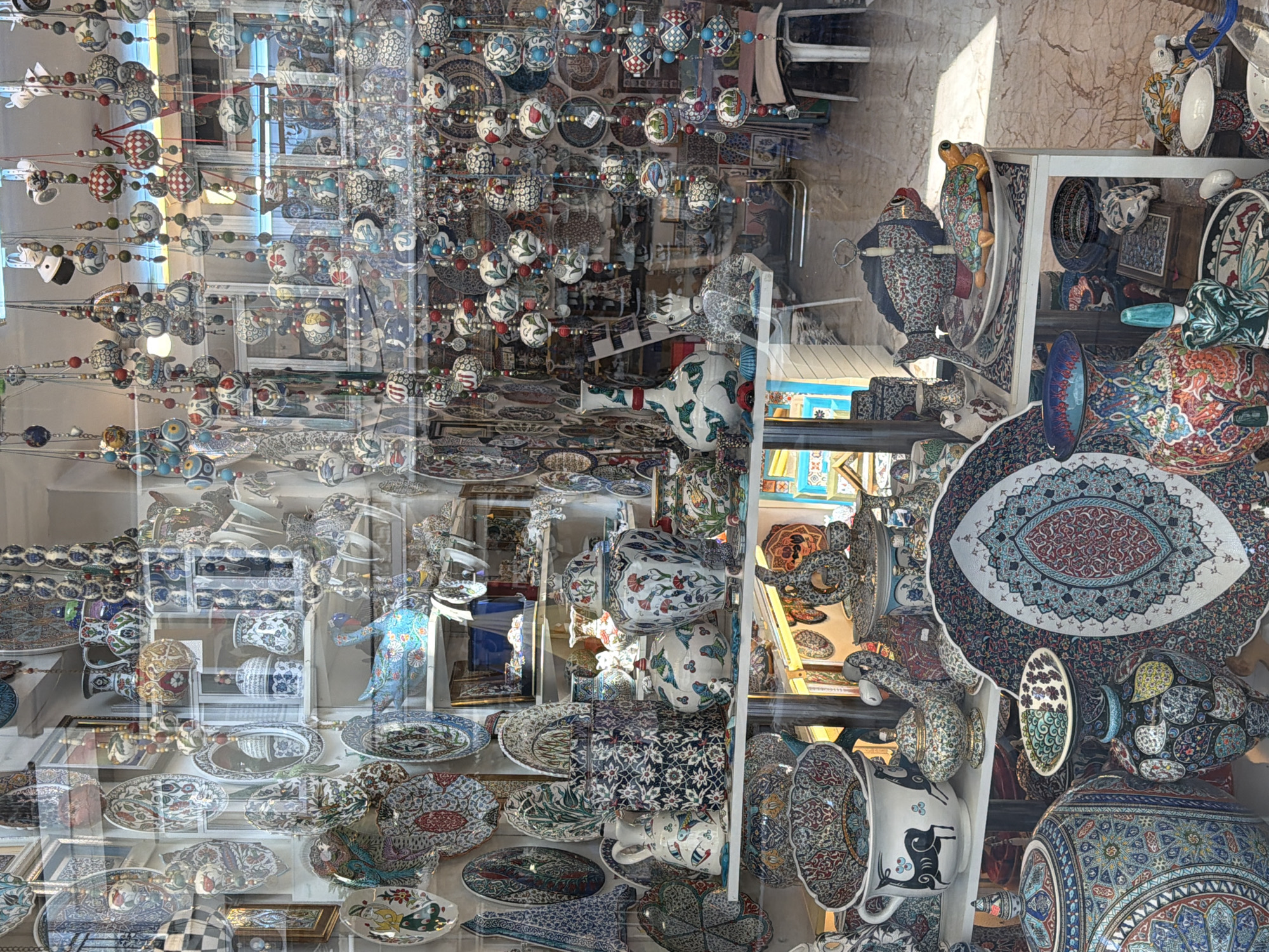 The image displays the interior of a shop filled with a diverse collection of decorative ceramics, likely a souvenir shop in Marmaris, Turkey. The items on display include intricately patterned plates, bowls, vases, and figurines, many featuring floral motifs and vibrant colors. The shop appears to be packed with merchandise, with shelves overflowing and decorative strings of ceramic beads hanging from the ceiling. Reflections of the outside environment, including sunlight and possibly other buildings, are visible on the shop window. The overall impression is one of a vibrant and abundant retail space specializing in handmade or artisanal ceramics, typical of tourist areas in Turkey.