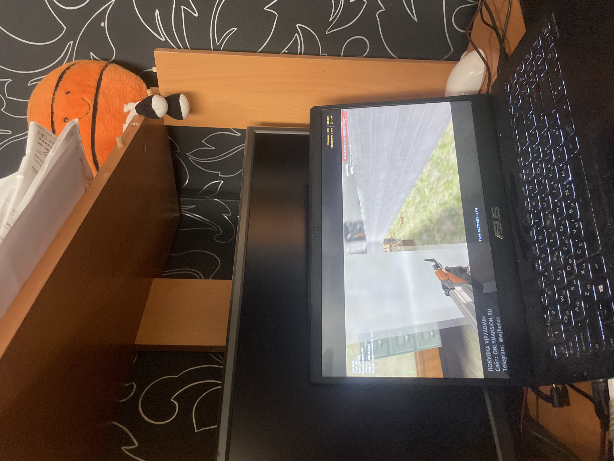 The image shows a cluttered desk area with a laptop displaying a video game, a computer monitor, and a plush basketball toy. The laptop is open and its keyboard is illuminated. The screen of the laptop displays what appears to be a first-person shooter game. In the upper right corner of the laptop screen, there are game statistics showing "Спецназ: 153" (Spetsnaz: 153) and "Террористы: 206" (Terrorists: 206) and a timer "1:18". Below the game on the laptop, there is text in Russian, reading "ПОКУПКА VIP/ADMIN", "Сайт: ONLYMANSION.RU", and "Telegram: @wjhenze". In the background, a wall with a black and white pattern is visible. A wooden shelf is mounted above the monitor. The plush basketball toy is orange with black stripes and a smiling face. A white computer mouse is visible to the right of the laptop. There is no indication of the city of Laishevo, Russia.