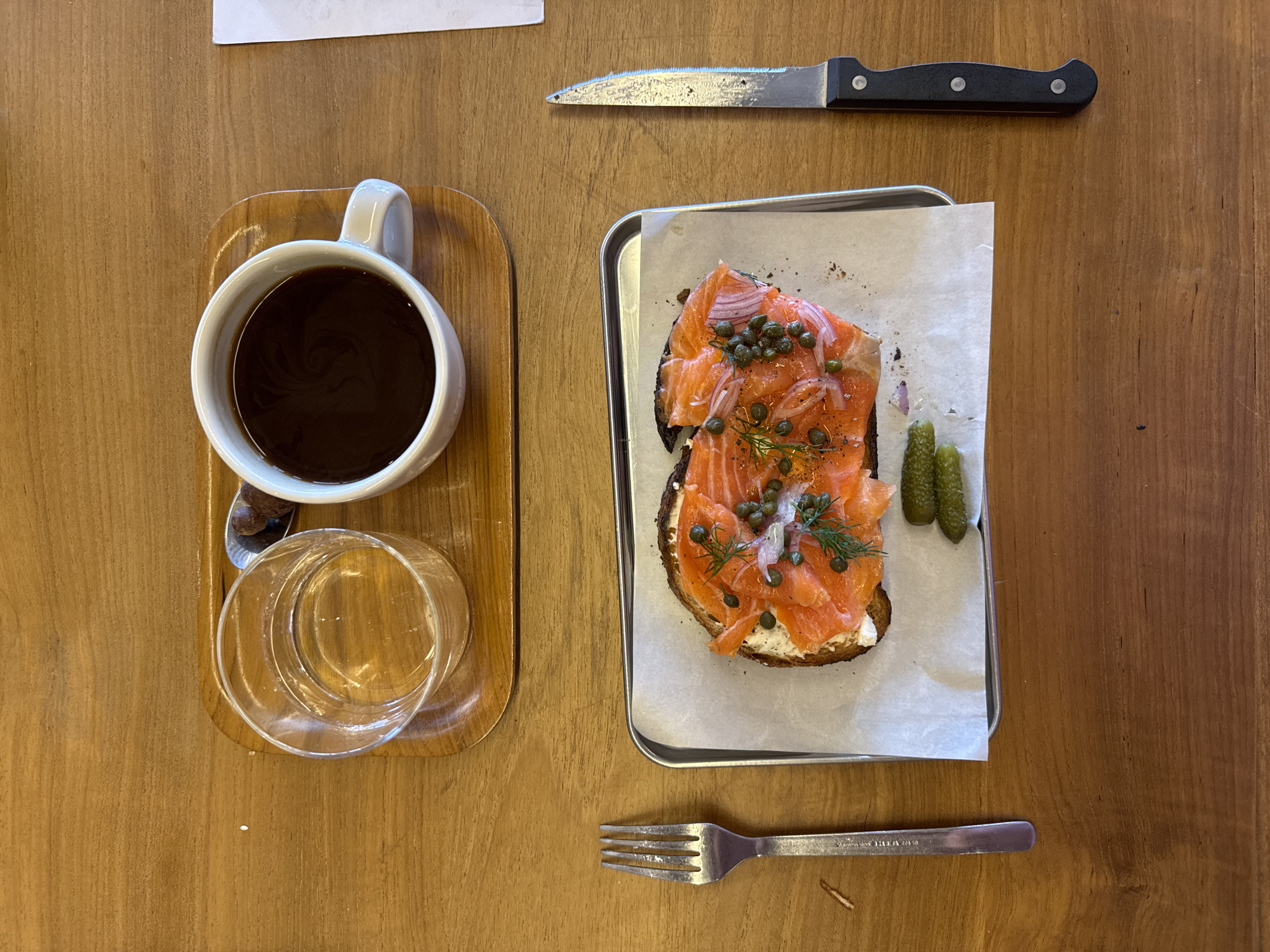 An overhead shot shows a wooden table setting with food and beverages. A wooden tray holds a white mug filled with dark coffee, a small spoon, and a clear glass of water. Next to the tray, a silver tray contains a slice of dark bread topped with cream cheese, smoked salmon, capers, and dill. Two small pickles lie beside the toast. A knife with a black handle is positioned above the silver tray, while a fork rests below. The scene suggests a cafe or restaurant setting, possibly for breakfast or brunch. The image does not provide sufficient information to confirm the location in North Kuta, Indonesia.