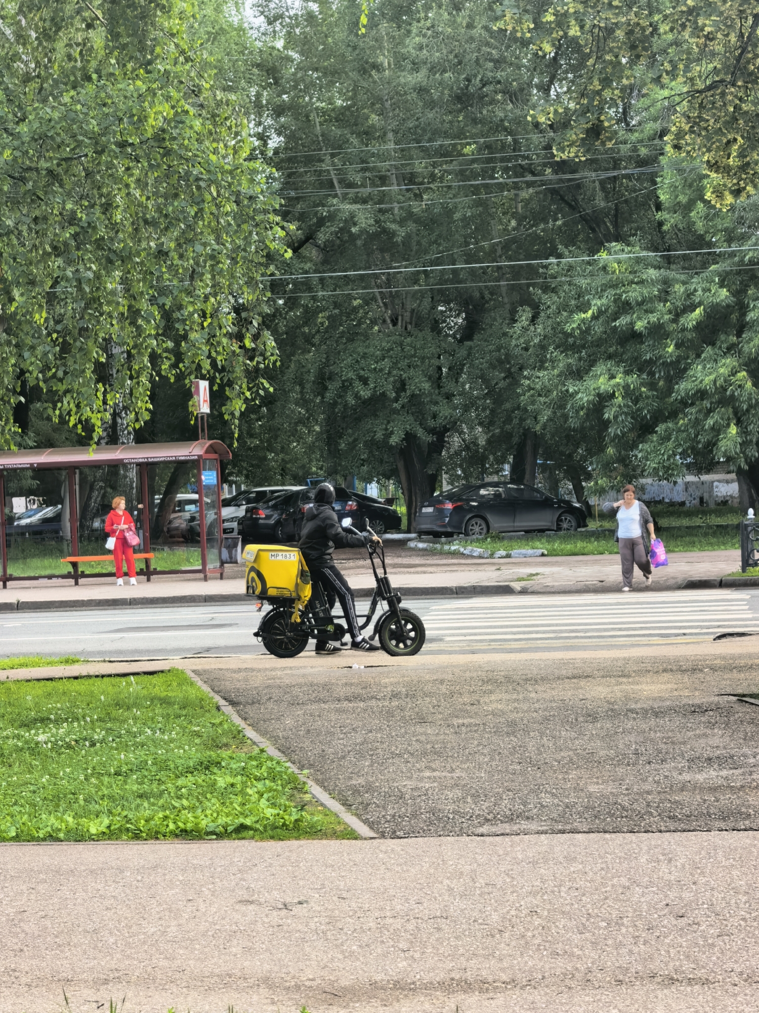 The image depicts a street scene in Sterlitamak, Russia, featuring a pedestrian crosswalk and a bus stop. A person on an electric scooter with a yellow delivery box is crossing the street. On the sidewalk, a woman with a shopping bag is walking across the crosswalk. A woman in a red tracksuit stands at the bus stop, which has Cyrillic signage indicating "ОСТАНОВКА БАШКИРСКАЯ ГИМНАЗИЯ" (Bus Stop Bashkir Gymnasium). Several parked cars are visible in the background, along with lush green trees and overhead power lines. The ground is a mix of asphalt and grass.