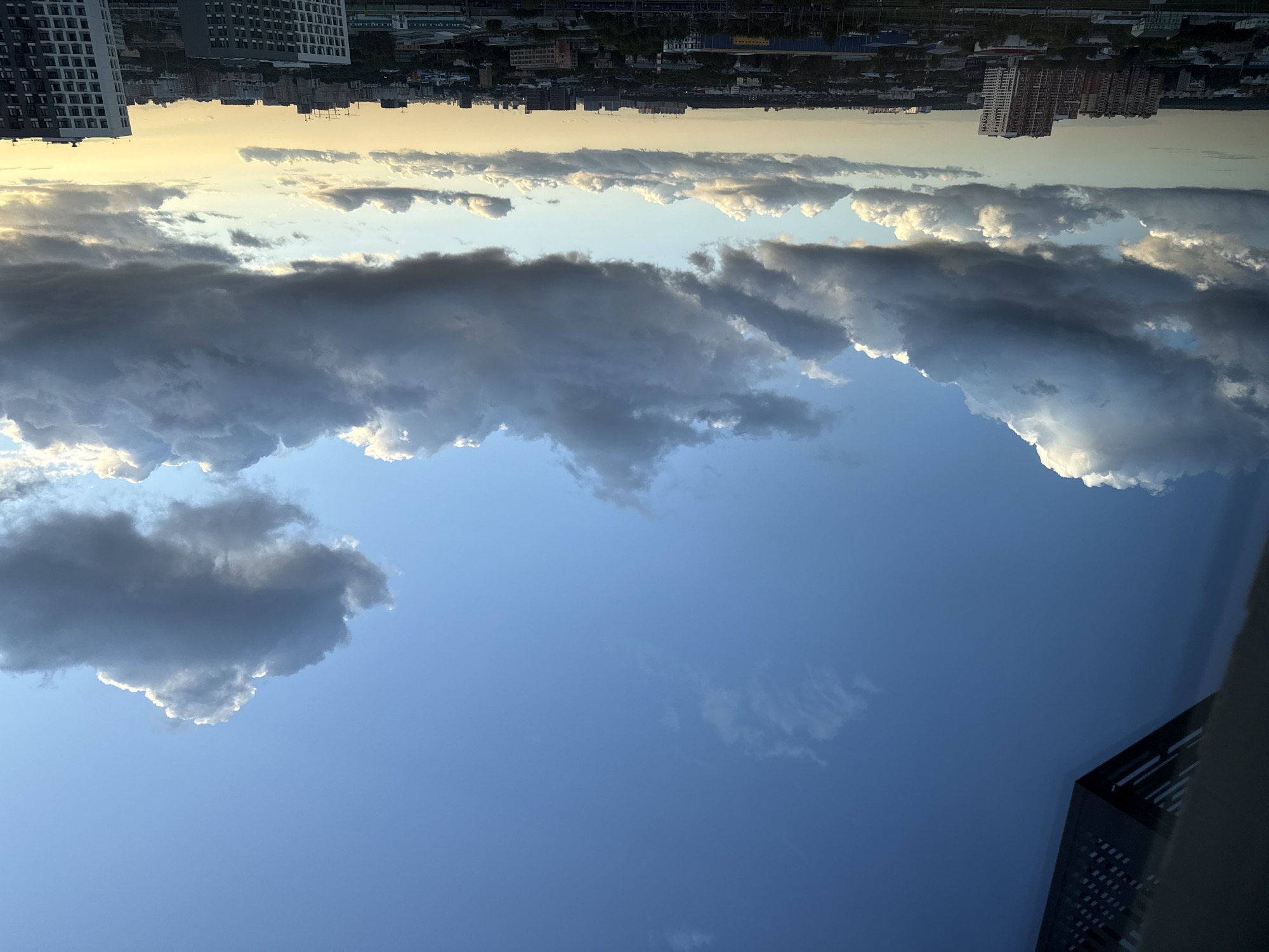 This is a medium-shot, eye-level photograph of a cityscape under a dynamic sky, likely taken from an elevated vantage point in Lyubertsy, Russia. The foreground is dominated by a large, dark cloud formation that occupies the upper half of the frame, with some scattered white cumulus clouds visible below it. The lower portion of the image showcases a sprawling urban landscape with numerous buildings of varying heights and architectural styles, including residential high-rises and what appear to be industrial structures. The setting sun casts a warm, golden glow on the horizon, visible between the clouds and the city. No people or active movements are discernible in the scene. The overall impression is one of a developing storm or an approaching sunset over a densely populated area.
