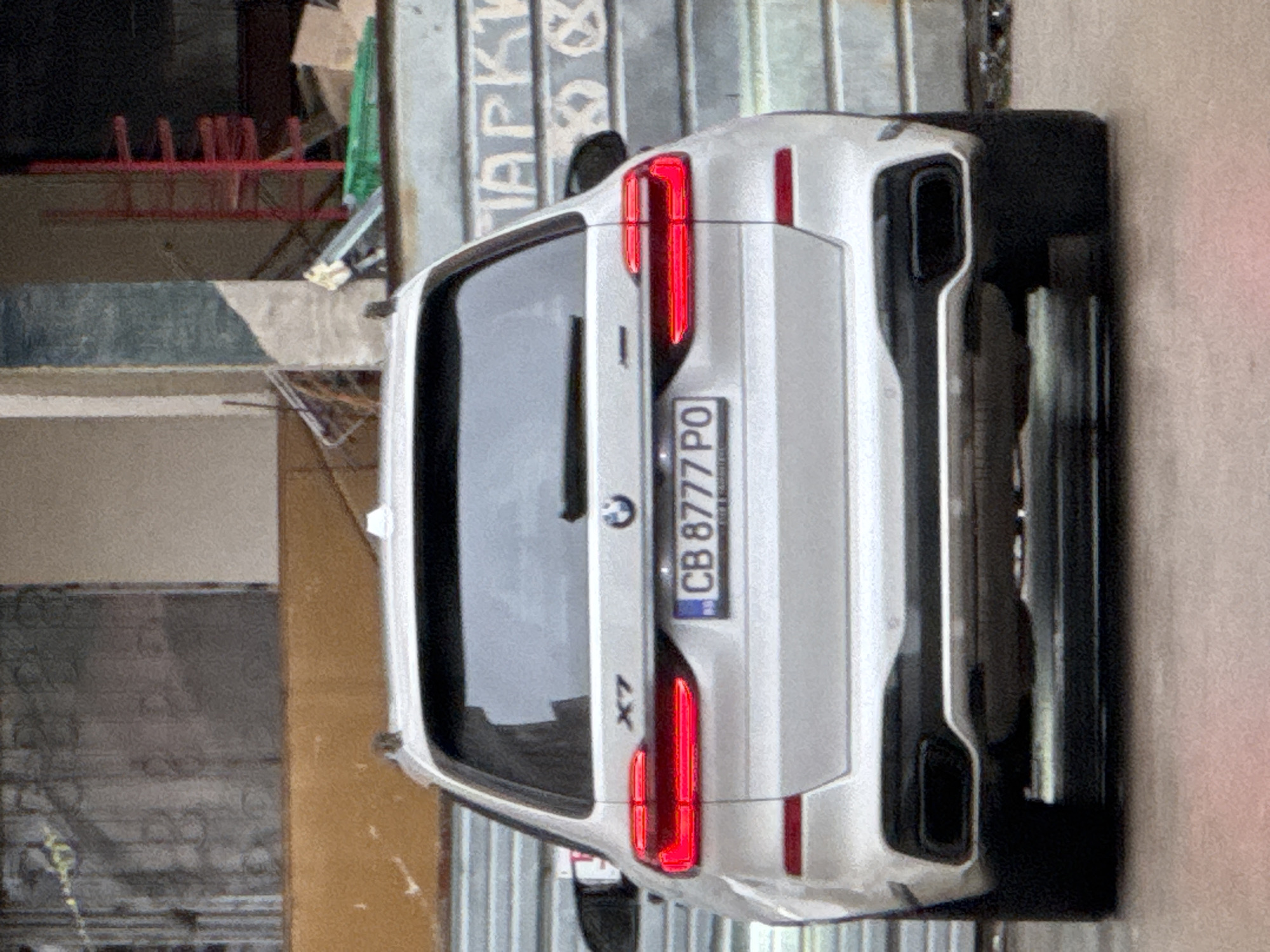The image shows the rear of a white BMW X7 with a Bulgarian license plate "CB 8777 PO". The background appears to be an urban environment, likely in Sofia, Bulgaria, as indicated by the license plate prefix "CB" which is used in Sofia. The surroundings include metal fencing with the word "Паркинг" (Parking) partially visible, indicating a parking area, and parts of other buildings. There are no people visible in the image. The car is parked, with its taillights showing.