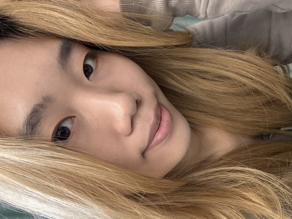 The scene is located in Nan'an District, China. It depicts a close-up, head-and-shoulders portrait of an East Asian female with a light skin tone. She possesses long, straight to wavy, honey-blonde hair that frames her face and extends over her shoulders. Her eyes are dark brown, and she has dark, well-defined eyebrows. She is looking directly into the camera with a subtle, closed-mouth smile, her naturally colored lips slightly upturned. Her head is tilted slightly to the right. She is wearing a light grey long-sleeved garment, partially visible on her right side. The background is shallowly focused, indicating an indoor setting with diffused lighting and a blurred, light blue-green surface or wall.