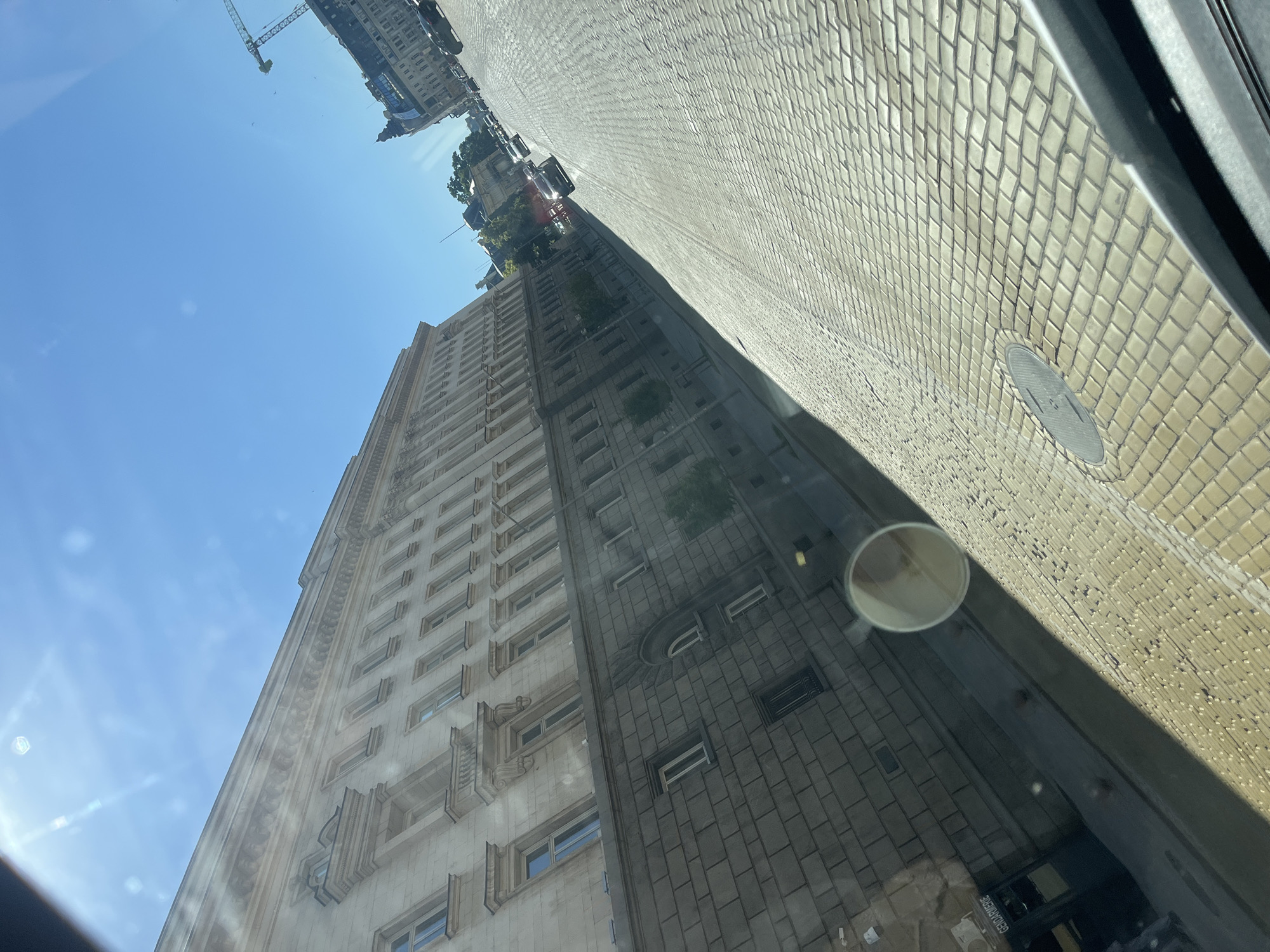 The image captures a view of a city street paved with cobblestones under a clear blue sky. On the left, a large, ornate building with multiple floors and windows dominates the scene. The building appears to be made of light-colored stone with detailed architectural elements. To the right of the building, the street slopes away, with several cars visible driving on it. In the distance, more buildings and a construction crane can be seen. The sun is shining brightly, casting shadows on the street. The perspective suggests the image was taken from a vehicle, possibly a bus or car, looking out a window.