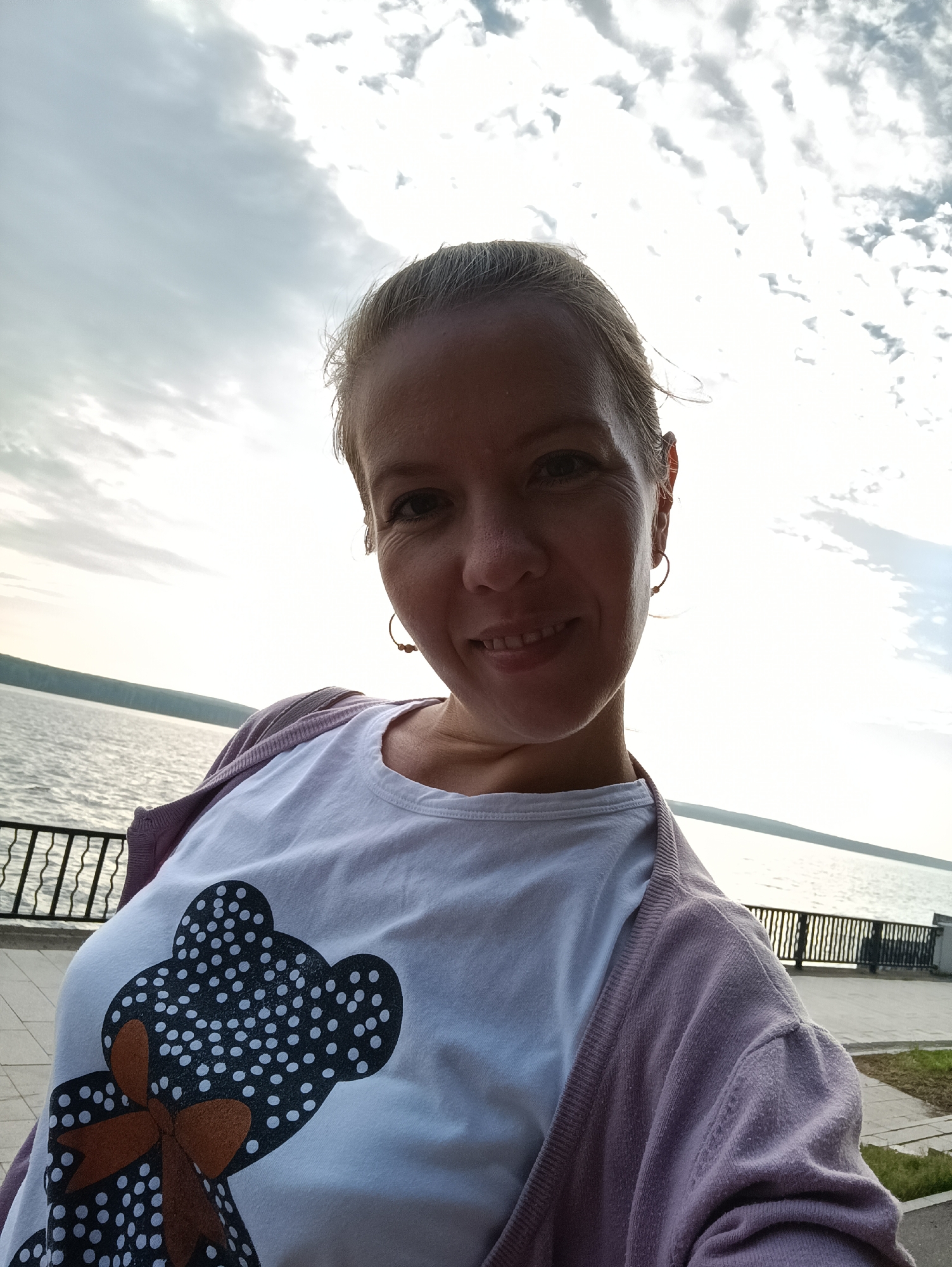 A woman with blonde hair pulled back poses for a selfie outdoors, smiling at the camera. She is wearing a white t-shirt with a black and white polka-dot teddy bear graphic and a light purple cardigan. She has small hoop earrings. The background shows a body of water, likely the Kama River, with a metal railing along a paved walkway and a distant treeline under a cloudy sky. The location is the embankment in the city of Chaikovsky, Russia.