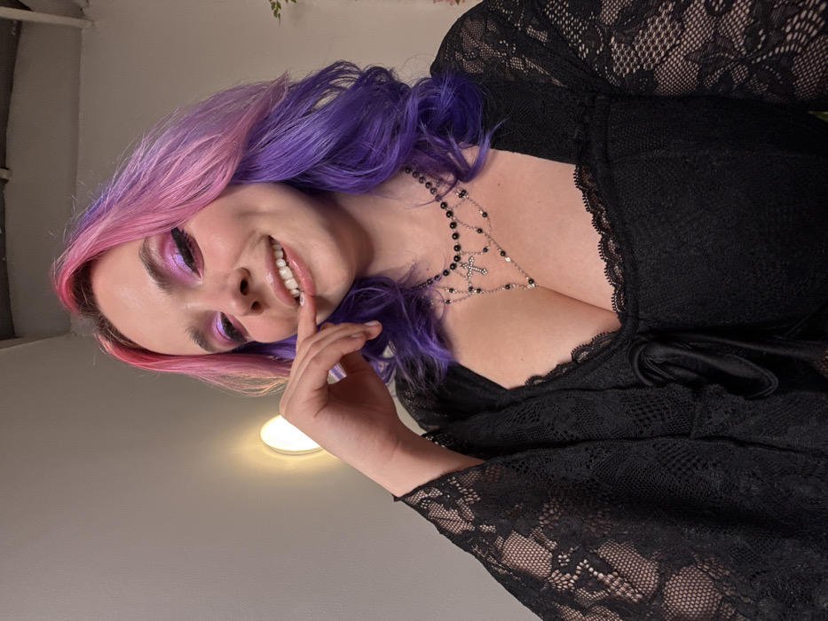 An indoor portrait displays a female subject in the City of Zagreb, Croatia. The subject has fair skin and medium-length, wavy hair dyed pink at the roots and fading into purple at the ends. She smiles, showing her upper teeth, and wears iridescent pink eyeshadow with eyeliner, and glossy lips. Her right hand is partially raised, with the index finger positioned near her mouth. She is dressed in a black lace top featuring a square neckline, long bell-like sleeves, and a central tie detail. A multi-layered necklace adorns her neck and chest, comprising strands of black beads and silver chains with a prominent silver cross pendant. The background consists of a plain light-colored wall with a circular wall-mounted light fixture visible in the upper left quadrant. A portion of green foliage is faintly discernible on the right side of the frame.