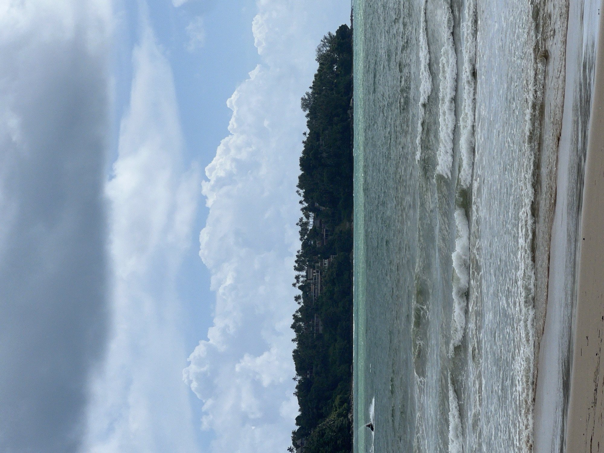 A person is jet skiing in the teal-colored ocean off the coast of Choeng Thale, Thailand. The ocean has moderate waves with white foam crests. In the background, a lush green hillside is covered with dense tropical vegetation and dotted with buildings, likely resorts or private residences, nestled among palm trees. The sky above is a mix of light blue and heavy, dark gray clouds, suggesting a partly cloudy or potentially stormy day. The foreground shows a sandy beach.
