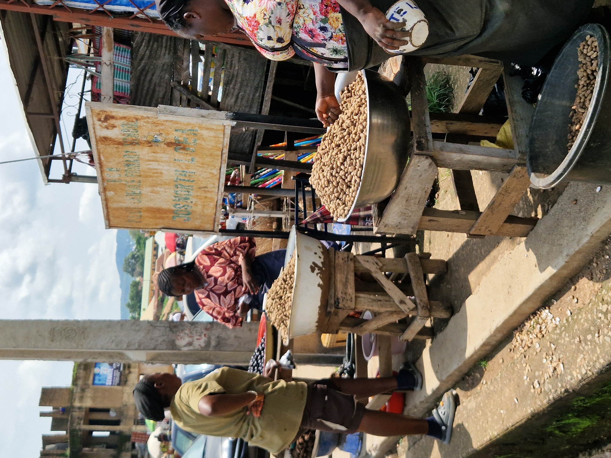 In Kabong, Nigeria, two women stand at a market stall. The woman on the left wears a green t-shirt and brown shorts, with a black headscarf. She is looking at the woman on the right. The woman on the right wears a vibrant, floral-patterned top and a dark apron. She is tending to a large bowl of peanuts. In front of her, on a wooden stand, is another bowl filled with peanuts, also for sale. Behind the women, market activity is visible, with other stalls and shoppers. A weathered sign with writing in English is mounted on a stand. The ground is paved with concrete and some areas are covered in dirt and debris. The scene is illuminated by daylight, suggesting it is daytime.